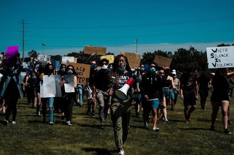 A large group of diverse protesters walking on a grassy field holding signs with messages about racial justice and social change, some wearing face masks, with a woman in the front using a megaphone.