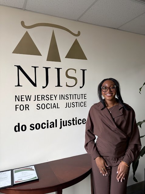 Woman standing in front of the New Jersey Institute for Social Justice sign at NJISJ with a table and framed certificate on it.