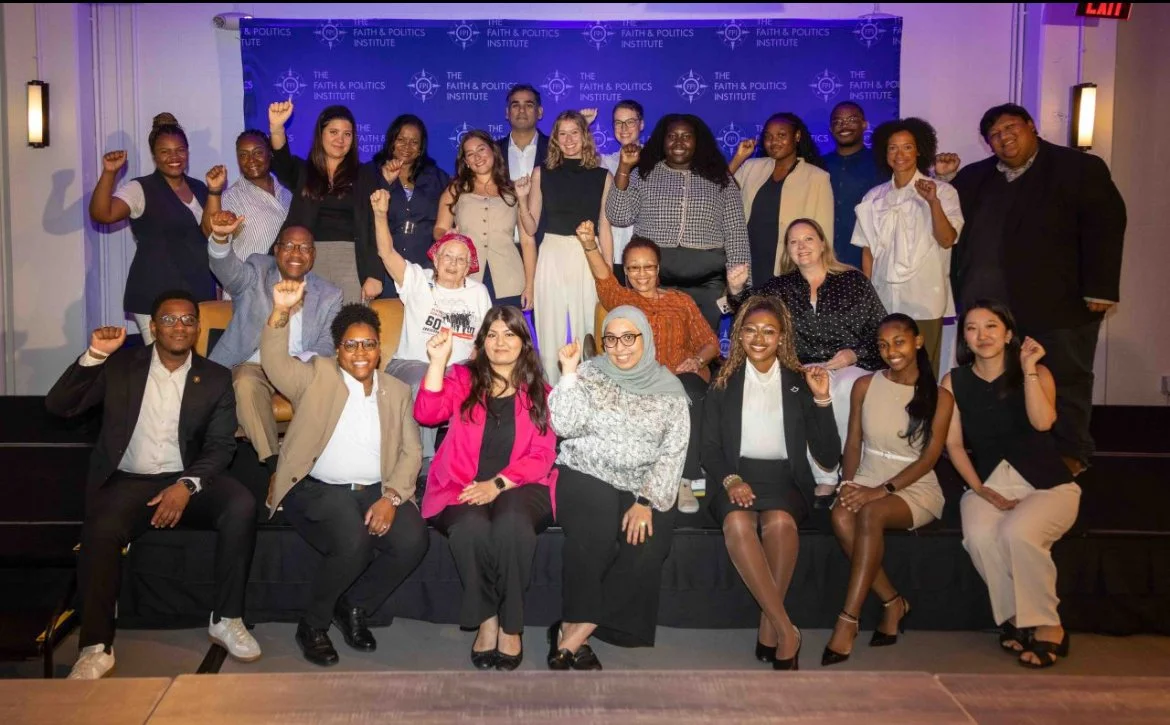 Group of diverse people in business and casual attire raising fists in celebration, standing and sitting on a stage with a blue backdrop that reads 'The Faith & Politics Institute'.