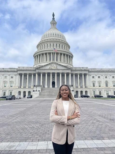 A young woman standing with arms crossed in front of the U.S. Capitol building on a cloudy day.