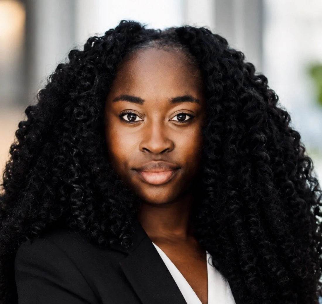 A portrait of a young Black woman with long, curly black hair, wearing a black blazer and white blouse, looking directly at the camera with a confident expression.