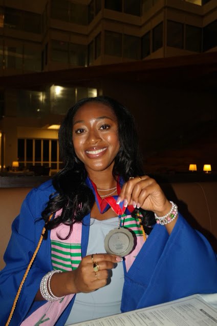 A young woman at a graduation celebration holding up a silver medal, wearing a blue graduation gown with pink and green accents, smiling and showing her diploma in a restaurant or banquet hall setting.