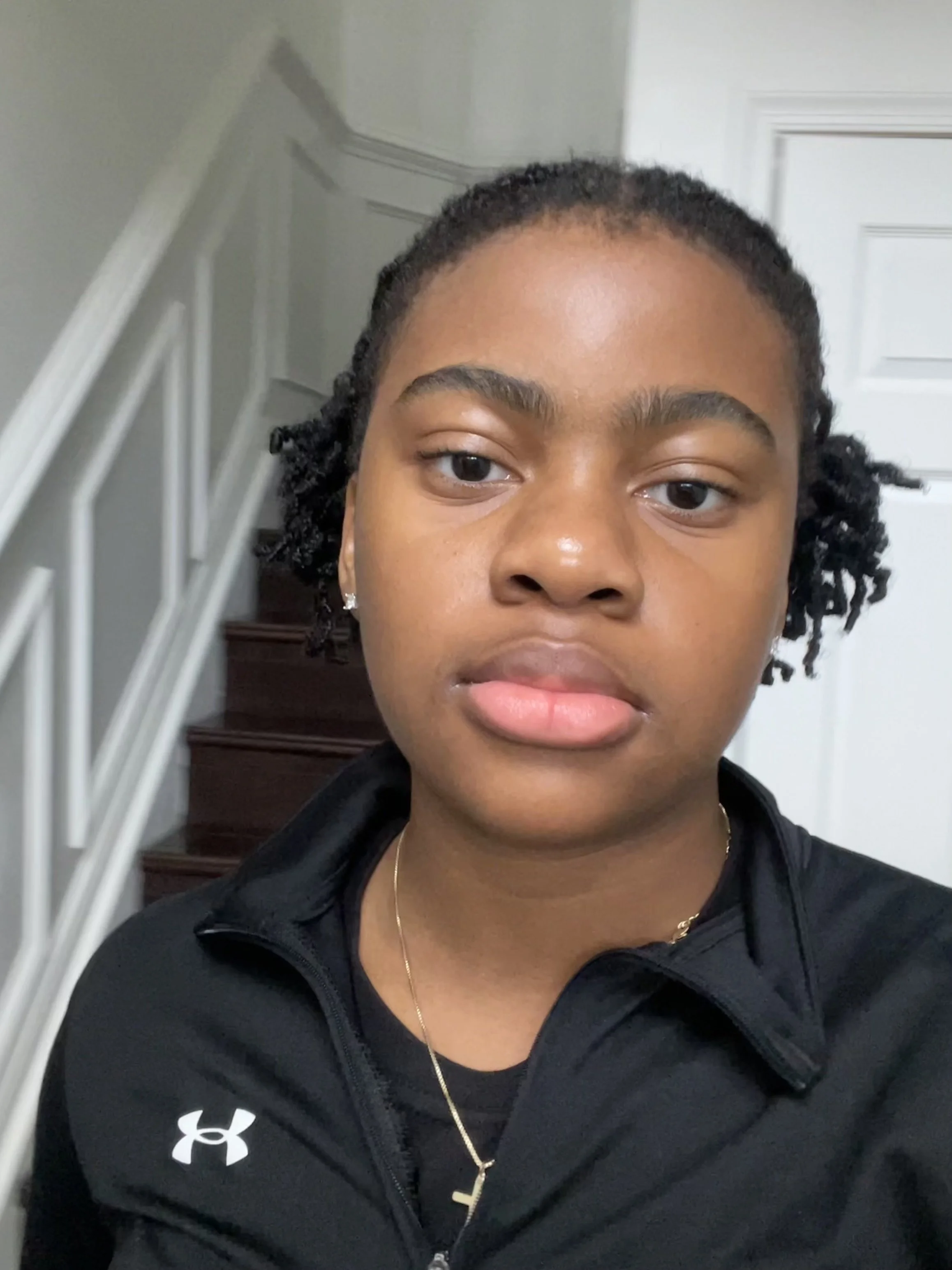 Close-up of a young woman with braided hairstyle wearing a black Under Armour jacket, gold necklace with initial Y, and small earrings, standing on a staircase near a white door.