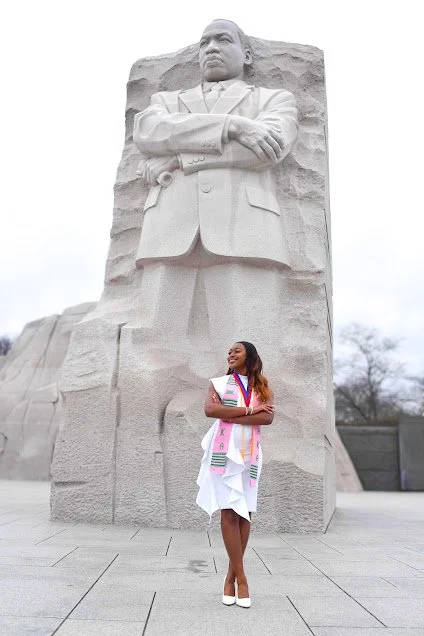 A woman in a white dress and colorful sash standing in front of the Martin Luther King Jr. Memorial statue of a large seated figure.