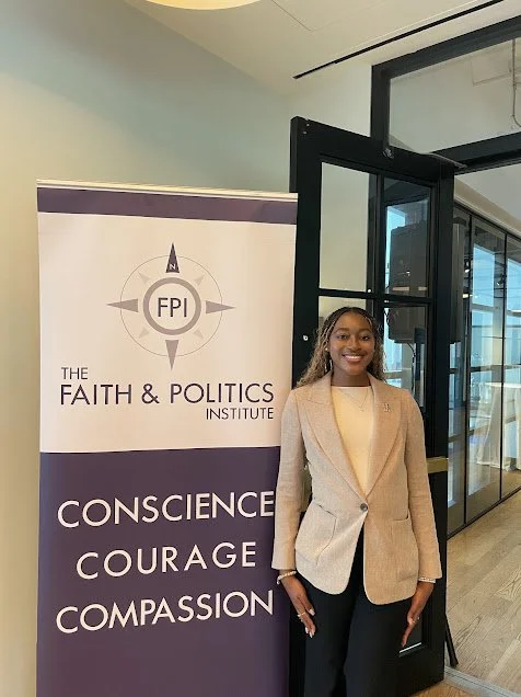 A woman standing next to a banner for The Faith & Politics Institute, which features a compass logo with 'FPI' in the center. The banner lists the words 'Conscience,' 'Courage,' and 'Compassion.'
