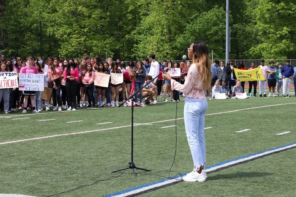 A young woman speaking at a microphone on a sports field during a protest or rally, with a group of students holding signs and banners in the background, surrounded by green trees.