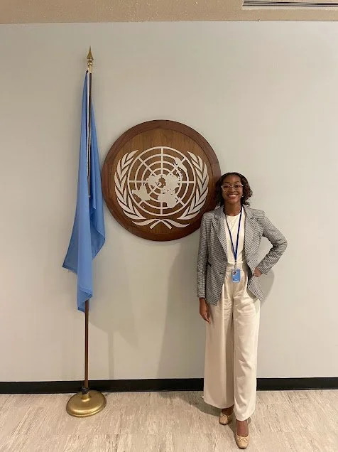 A woman standing in front of a United Nations emblem on a wall, with a UN flag on a stand to her left, smiling at the camera.