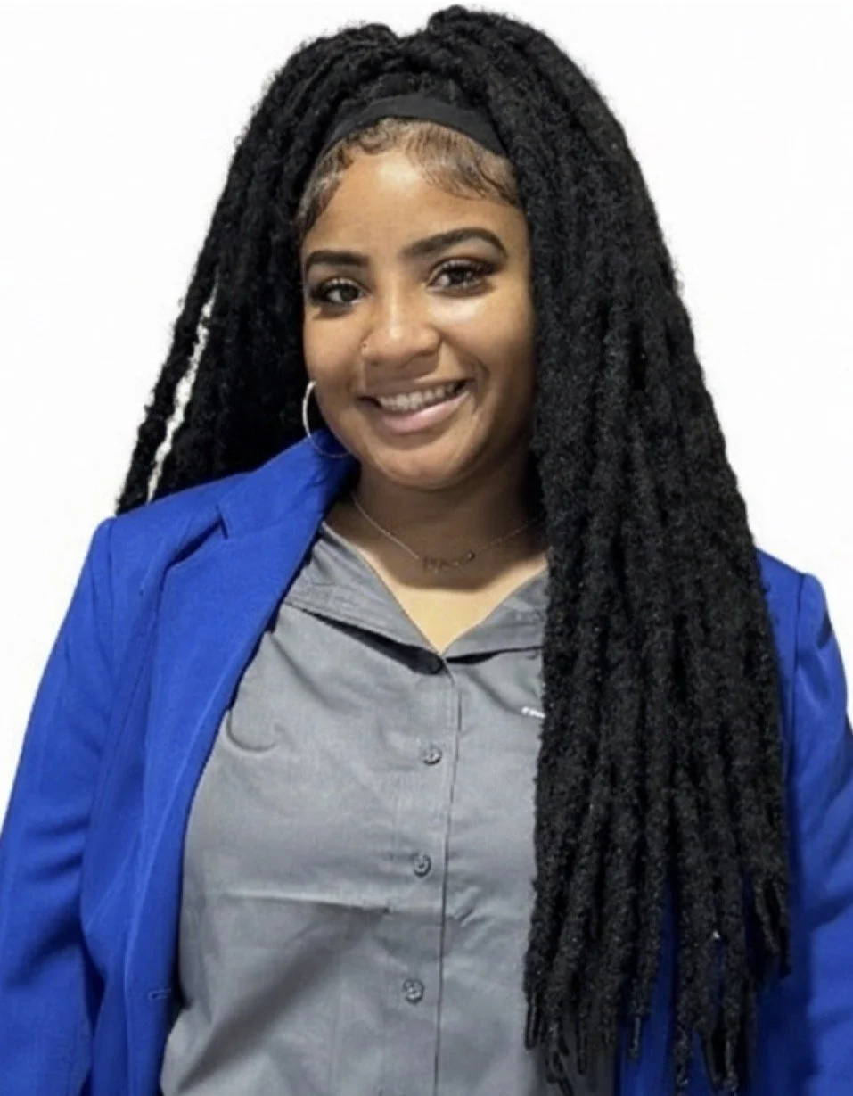 A woman with long black dreadlocks wearing a gray shirt and a blue blazer, smiling at the camera.
