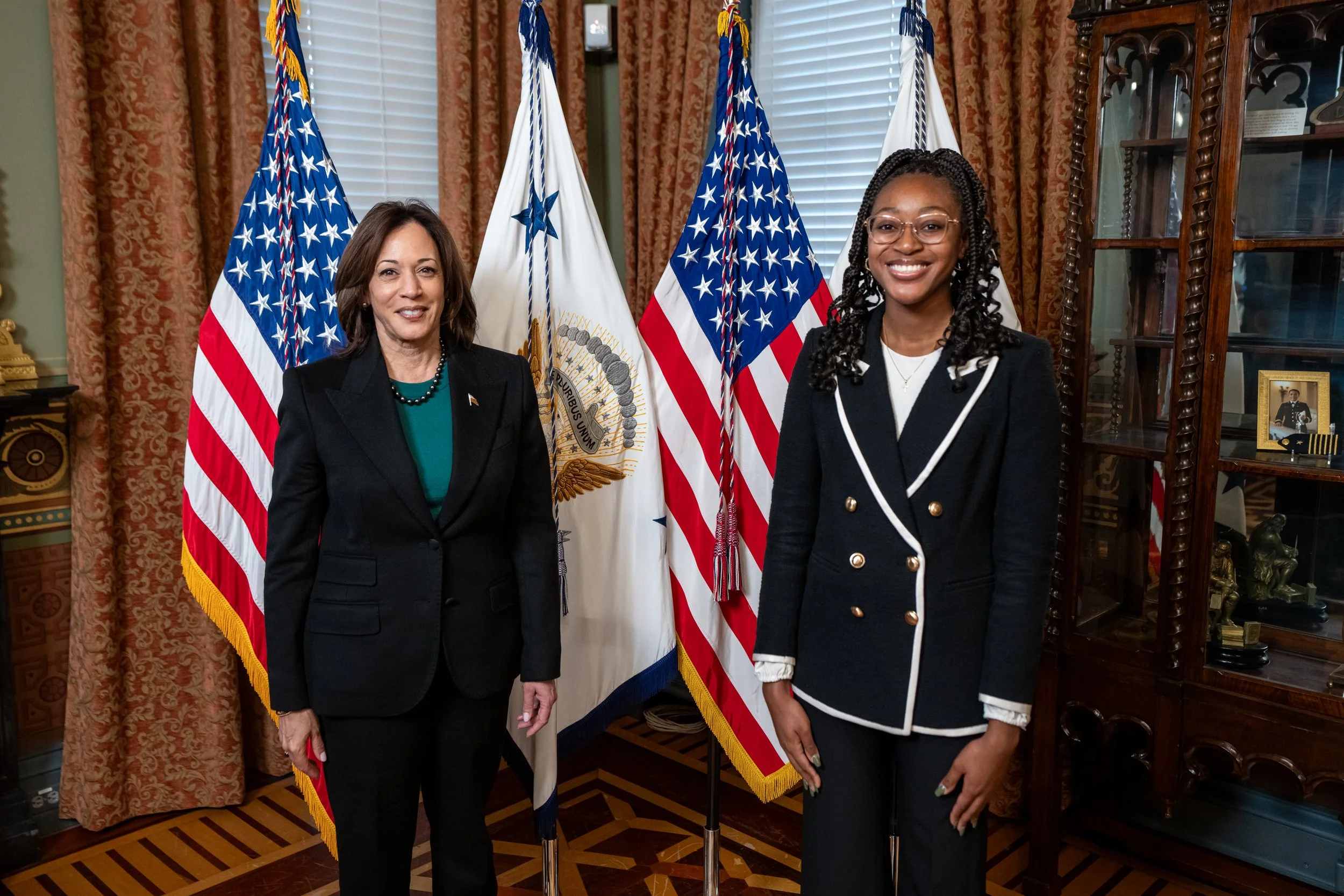 Two women standing side by side in a room with five American flags and a white flag between them, surrounded by antique wooden furniture and red and gold curtains.