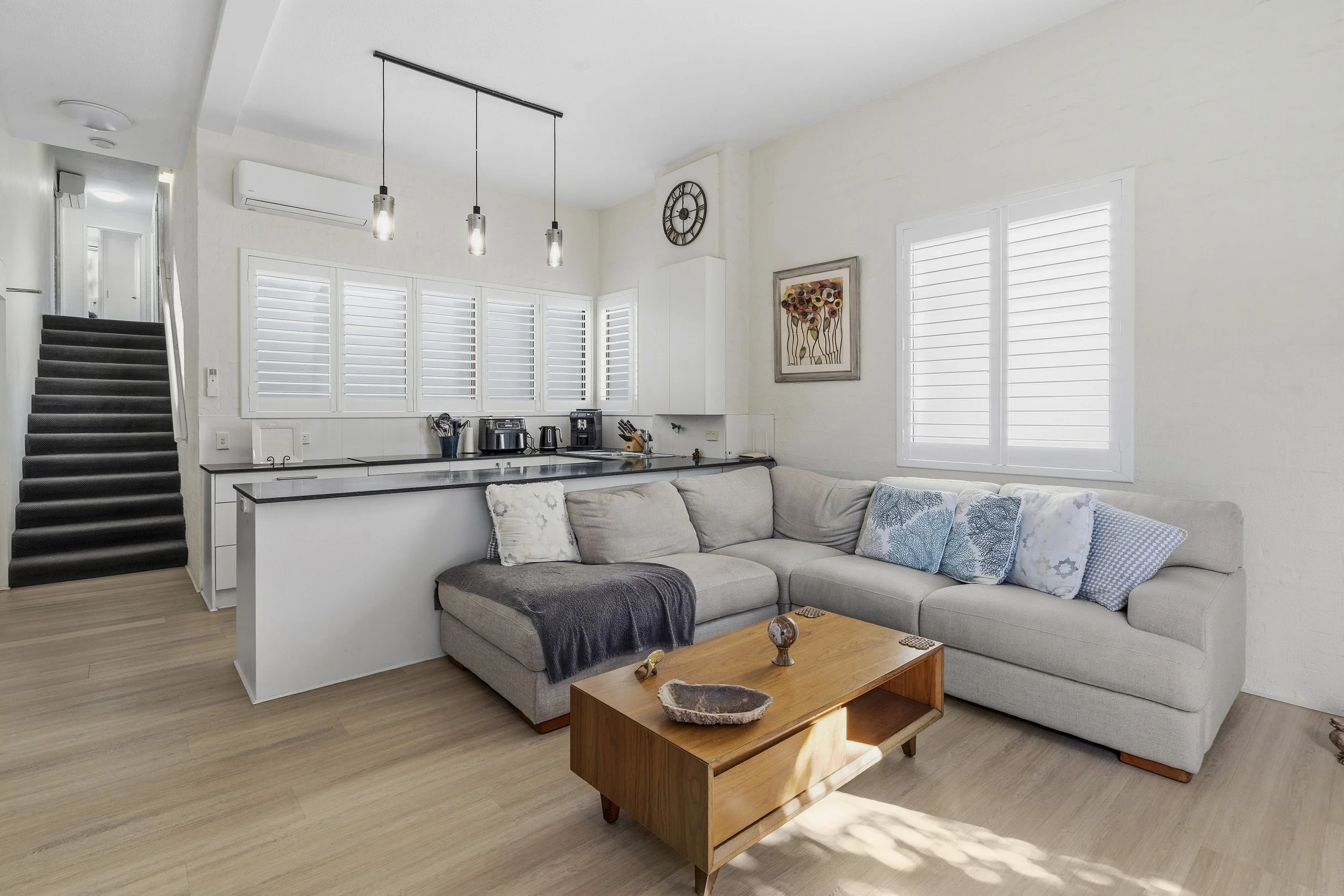 A modern living room with a beige L-shaped sofa, decorative pillows, and a wooden coffee table. Behind the sofa are large windows with white shutters, and the kitchen area with hanging pendant lights, countertop appliances, and a window with shutters