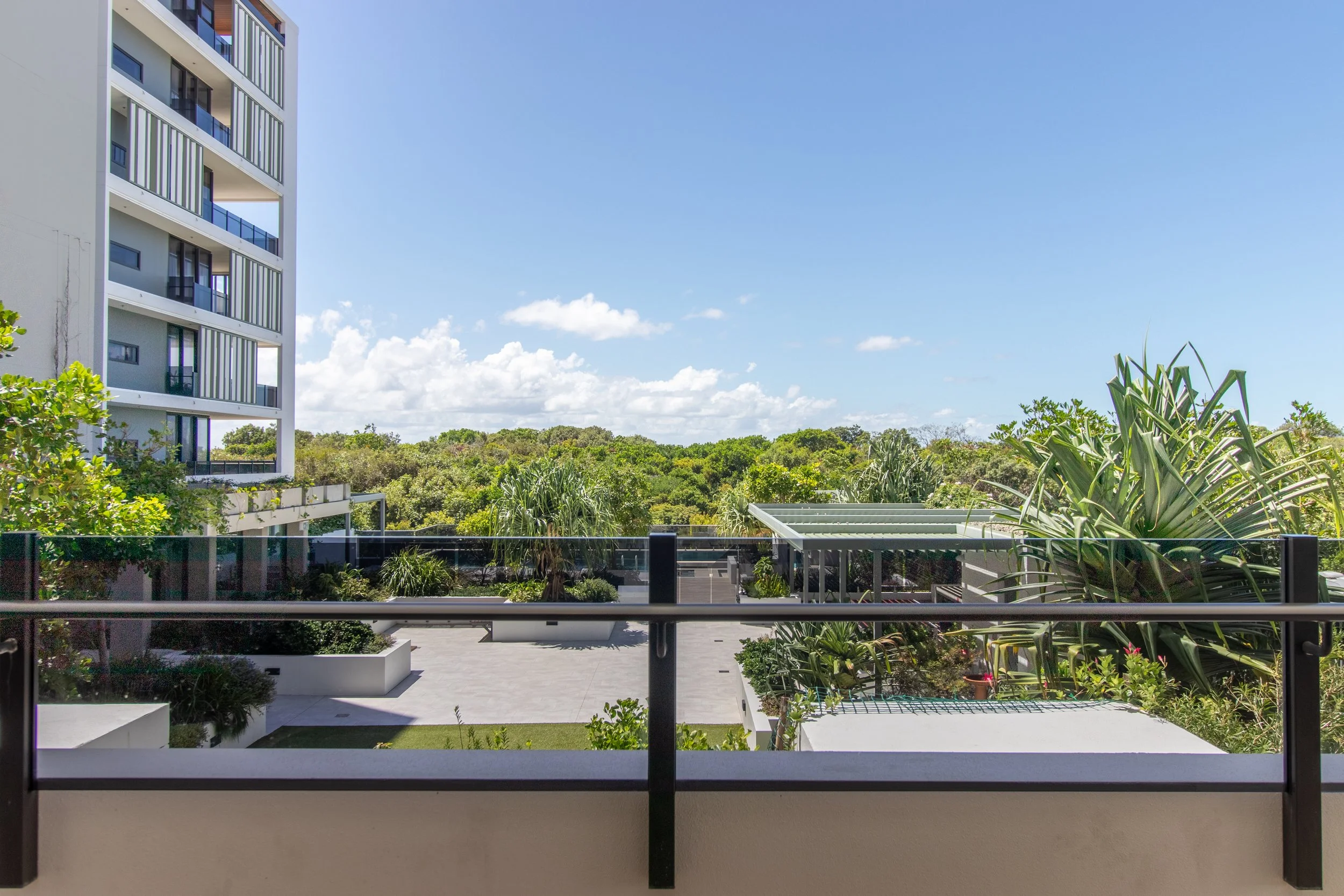 View from a balcony overlooking a parking lot, lush green trees, and a mostly clear sky with a few clouds.