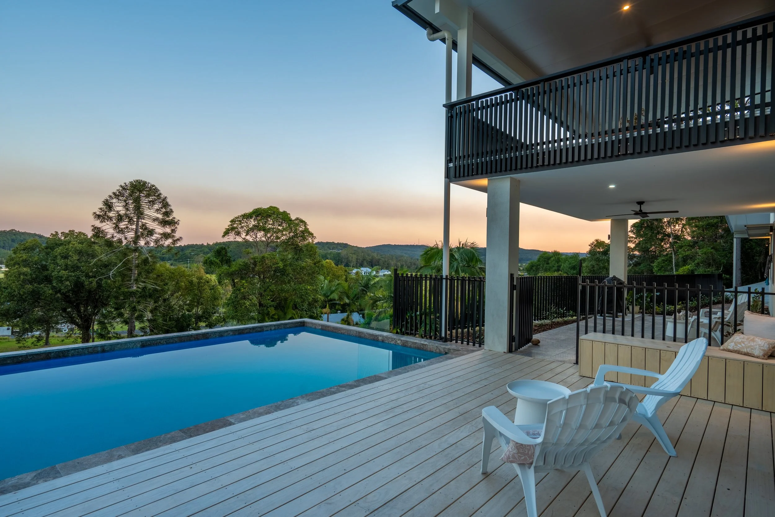 A modern house balcony overlooking a scenic landscape with trees and hills during sunset. The balcony features a swimming pool, white deck chairs, and a privacy fence.
