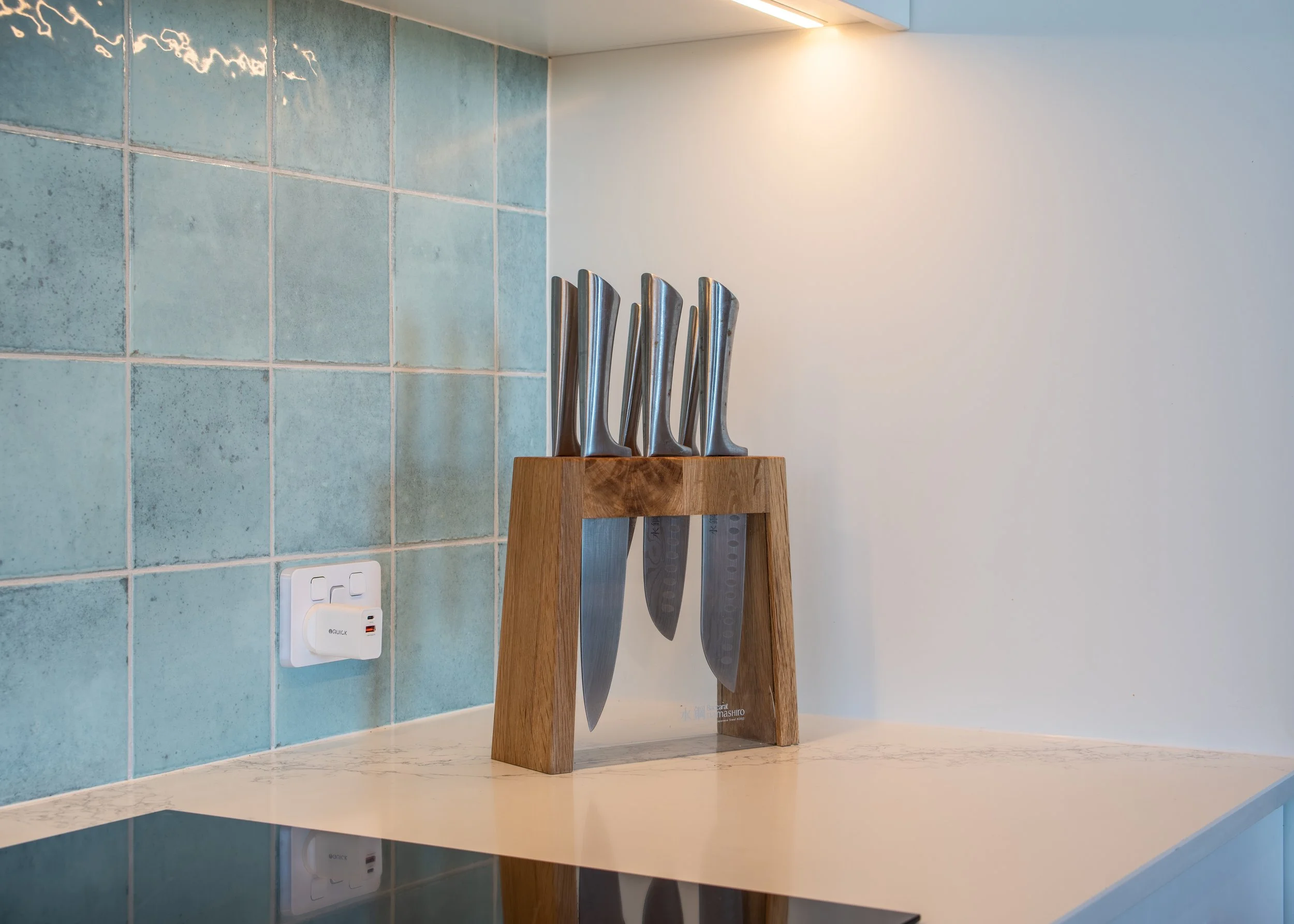 Seven kitchen knives placed on a wooden knife holder on a marble countertop, with a tiled wall and electrical outlets in the background.