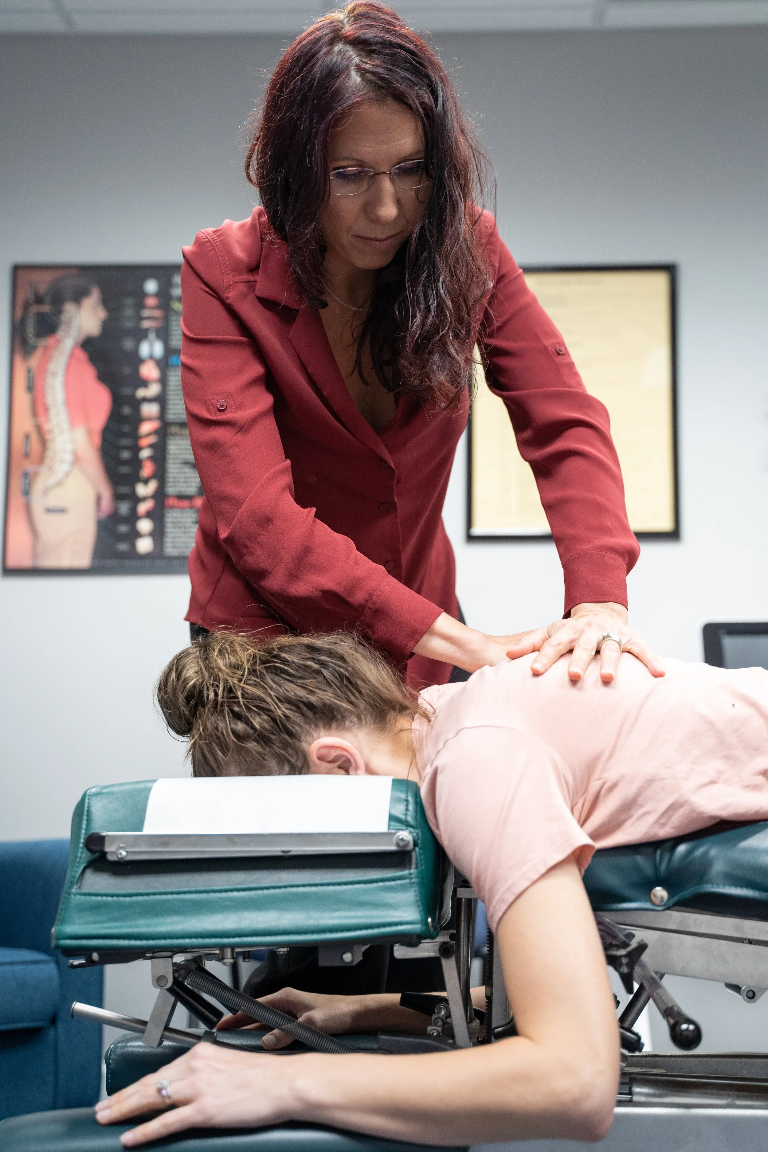 A woman with glasses and long dark hair wearing a red shirt performs a chiropractic adjustment on a patient lying face down on a chiropractic table. The room has medical charts on the walls.