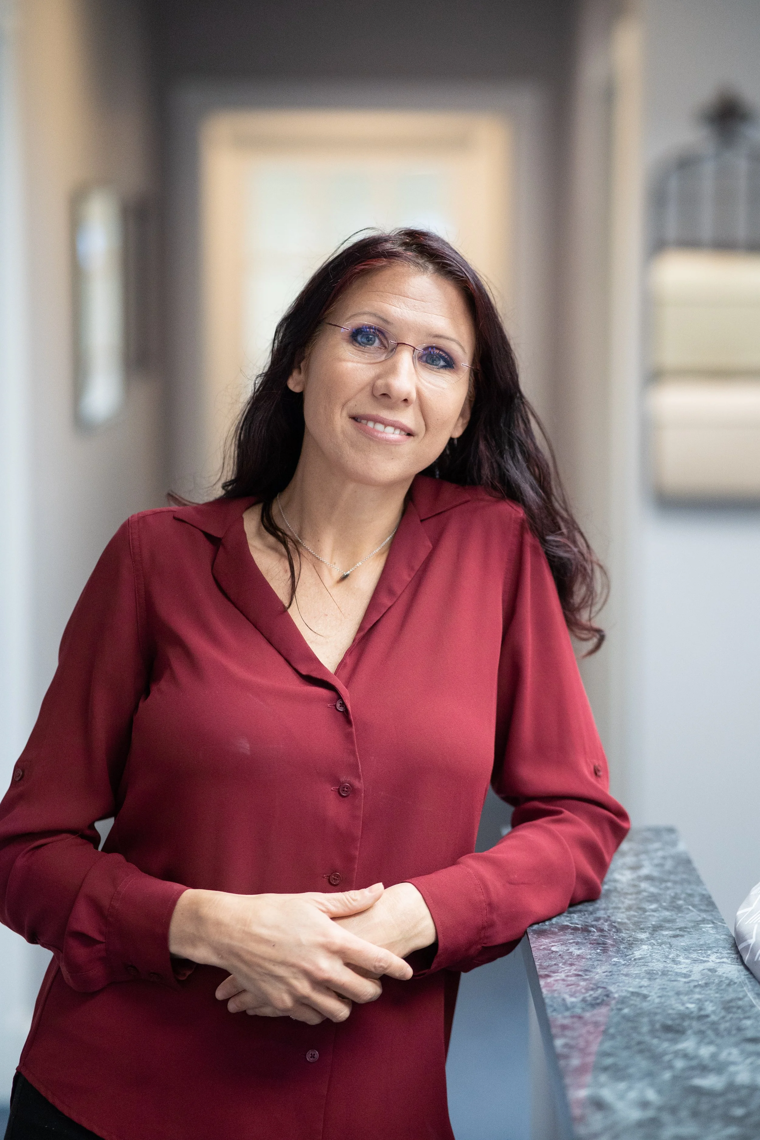 A woman with dark brown hair and glasses wearing a red blouse standing at a granite counter in a modern kitchen.