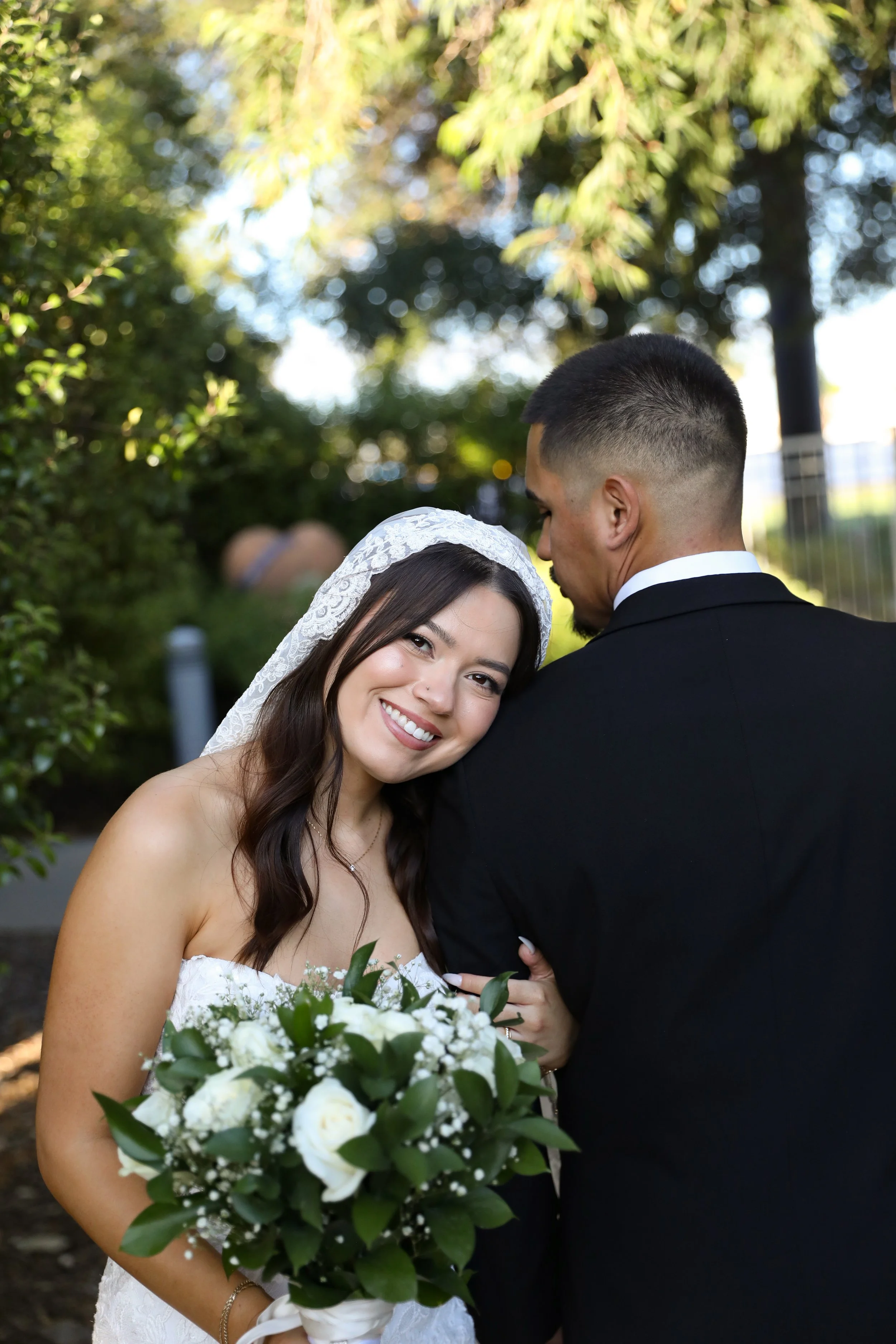 A bride with long dark hair wearing a white lace head veil, smiling and holding a bouquet of white flowers and green leaves, standing close to a groom with short dark hair, dressed in a black suit, outdoors with green trees and sunlight in the background.