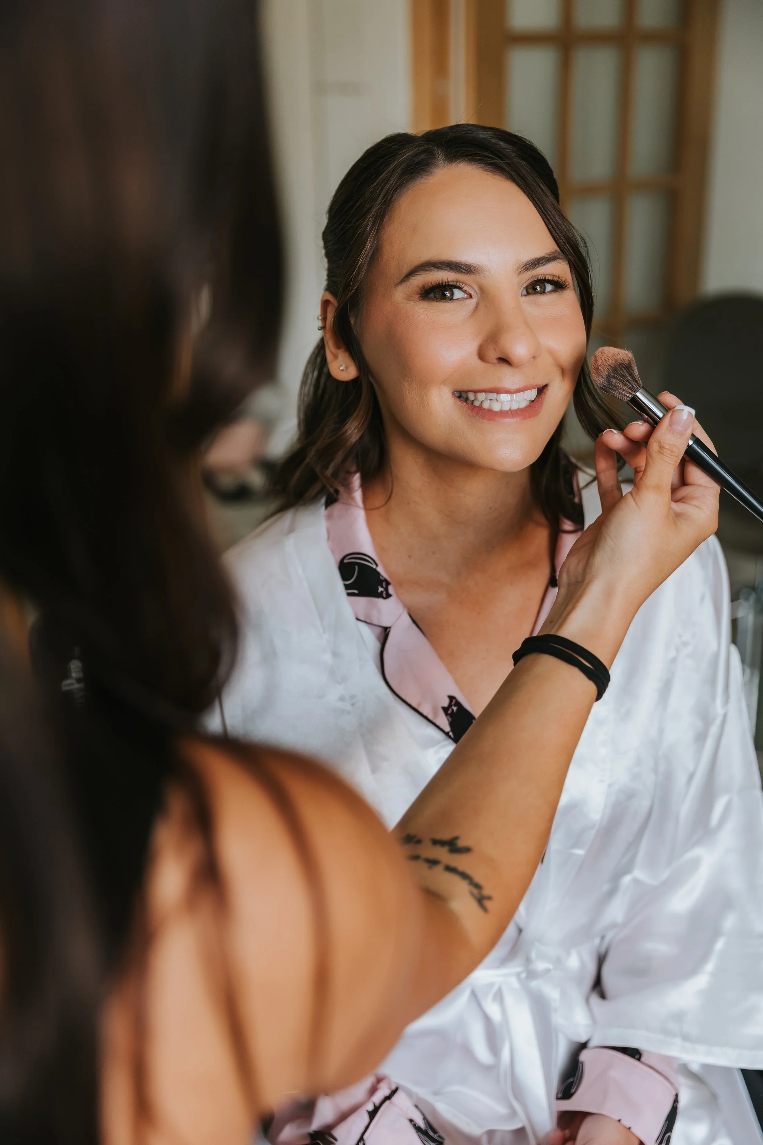 A woman smiling while someone applies makeup with a brush to her face.