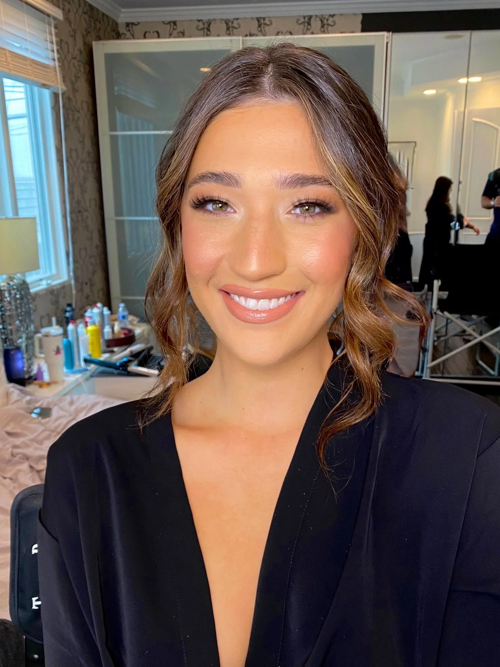 Close-up of a woman with wavy brown hair smiling, wearing a black top, in a room with windows, mirror, and various hair and makeup tools in the background.