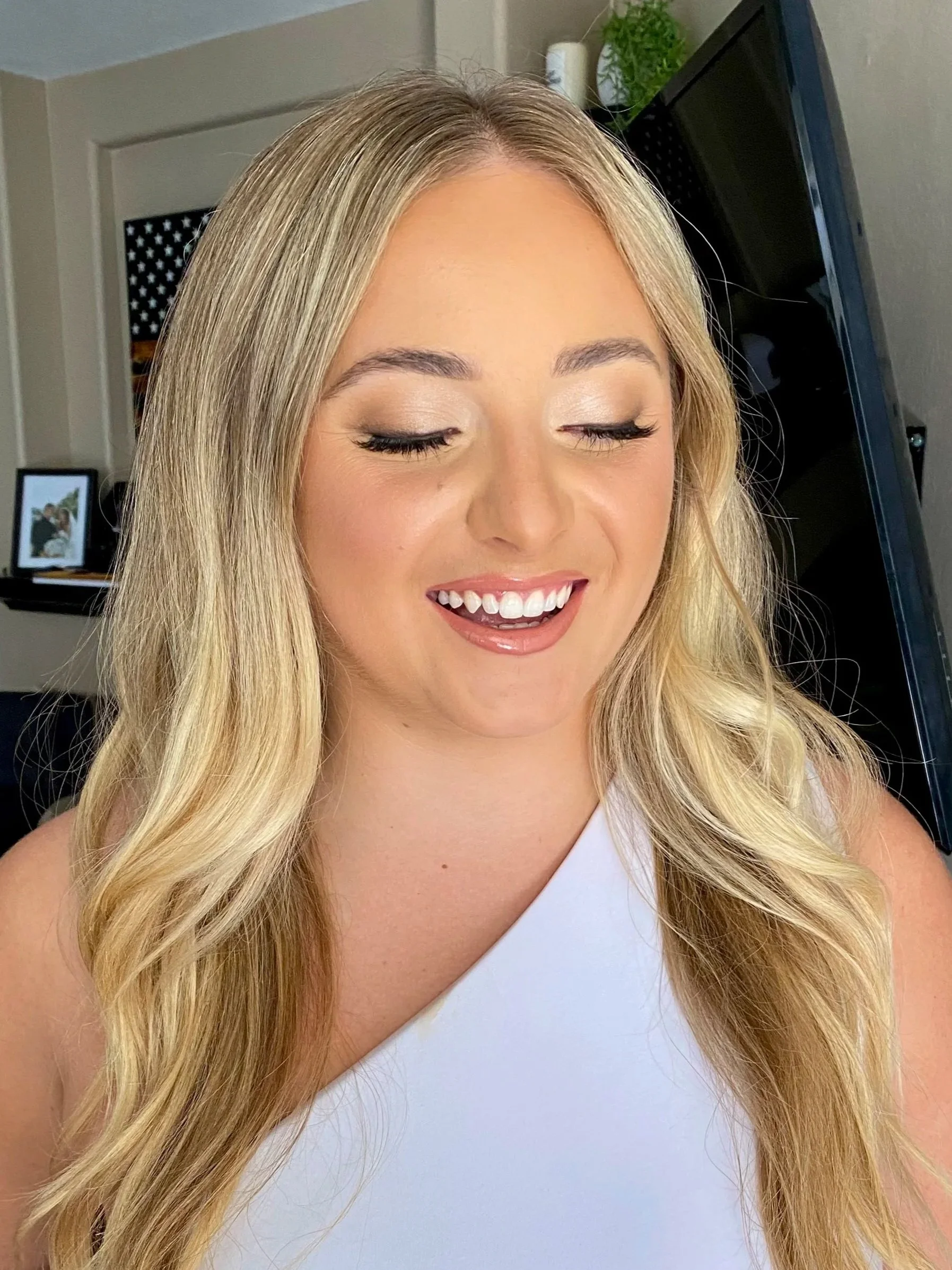 Close-up of a smiling woman with long blonde hair, closed eyes, and wearing a white sleeveless top, in a room with a computer, framed photo, and green plant in the background.