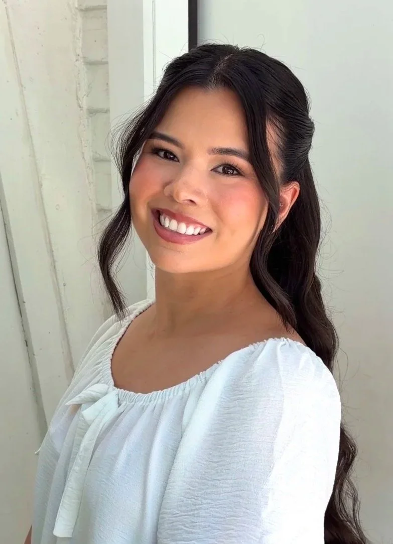 A woman with long dark hair smiling and wearing a white top, standing near a white wall and a window.