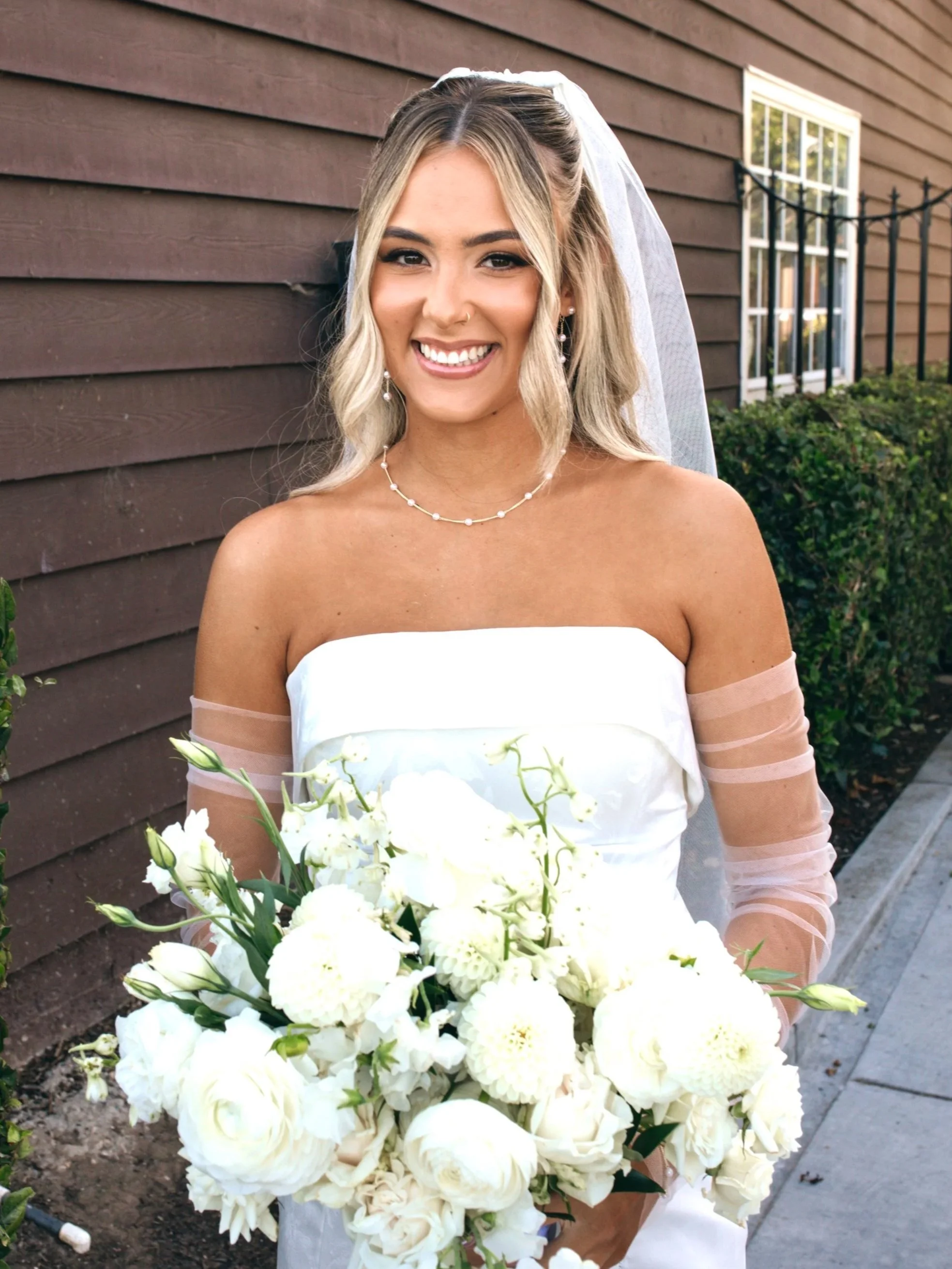 A smiling woman in a strapless white wedding dress holding a large bouquet of white flowers, standing outdoors near a wooden building.