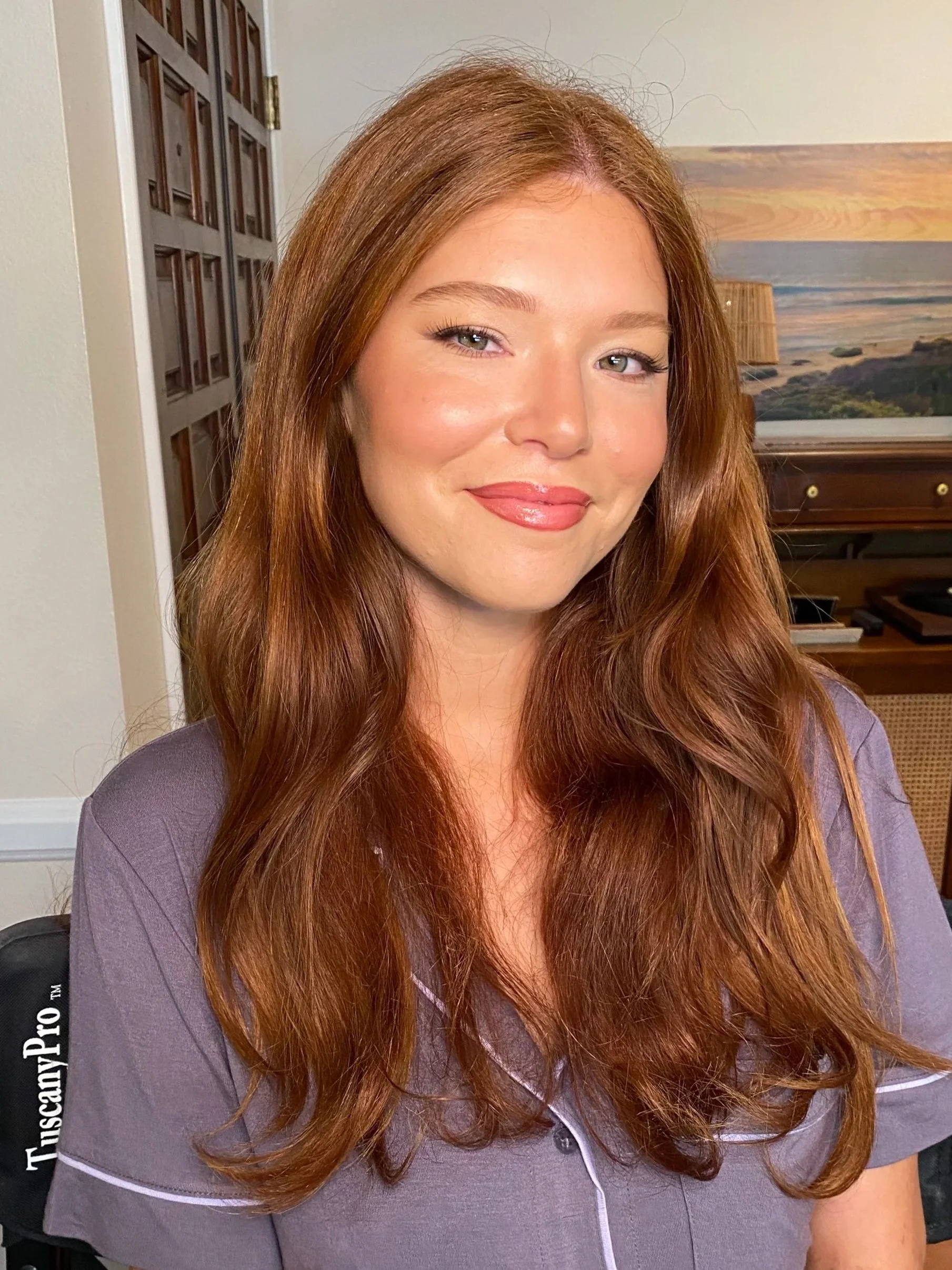 Close-up of a woman with long, wavy auburn hair, fair skin, and light eyes, wearing a purple top, smiling softly inside a room with a landscape painting and wooden furniture in the background.