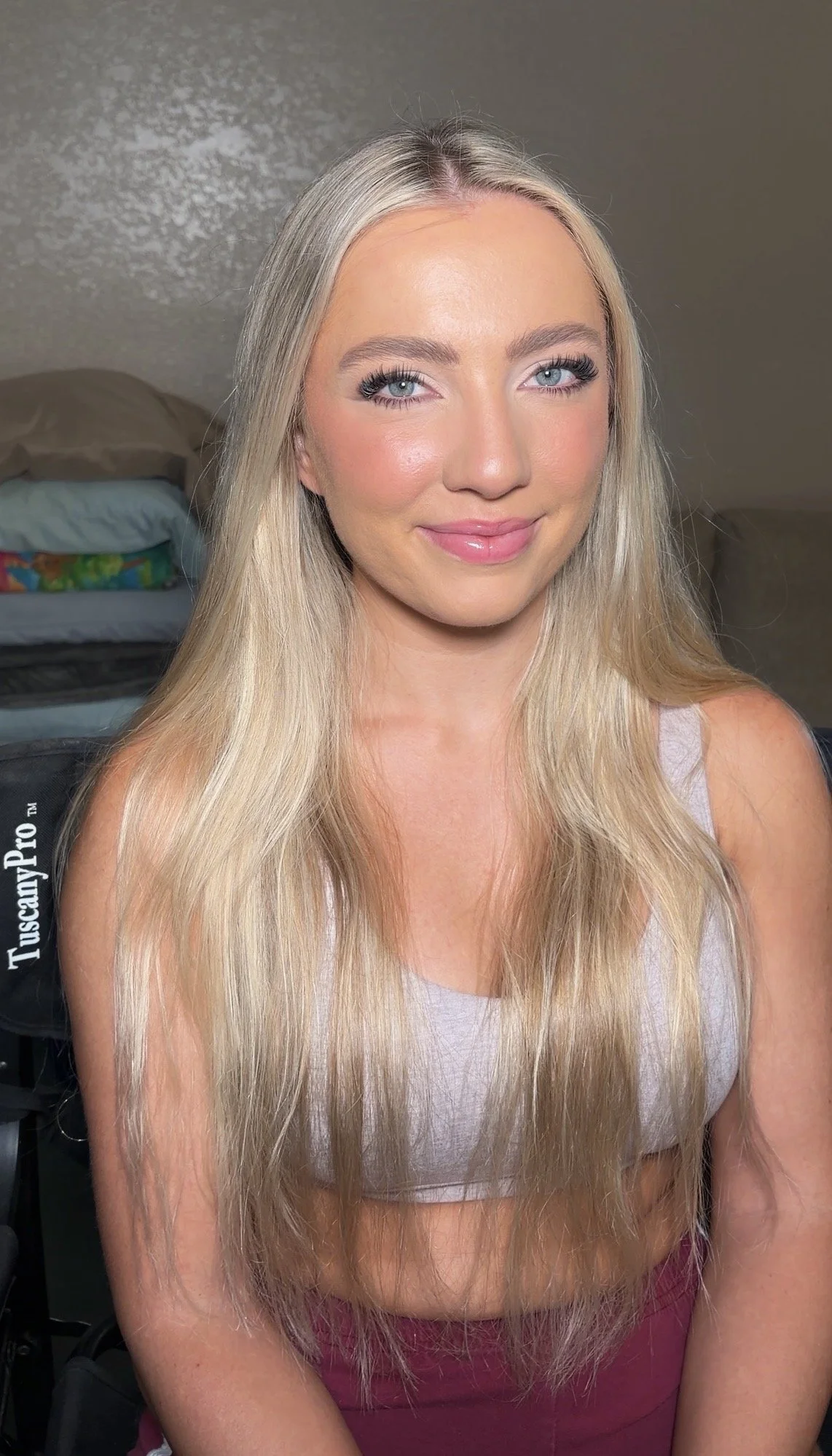 Close-up portrait of a young woman with long, blonde hair, blue eyes, and a gentle smile, sitting indoors with a neutral background.