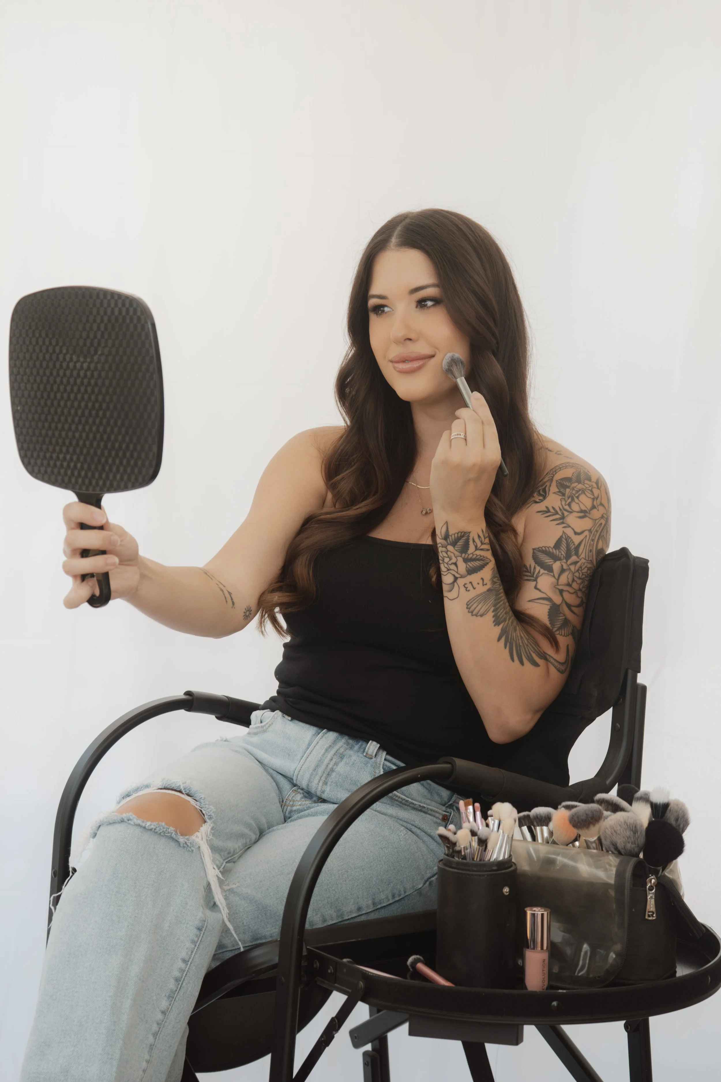 A woman with long brown hair and tattoos on her arms is sitting in a makeup chair, applying blush with a makeup brush while looking into a mirror. She is wearing a black tank top and ripped light jeans, with various makeup tools and products organized in a black makeup trolley in front of her. The background is plain white.