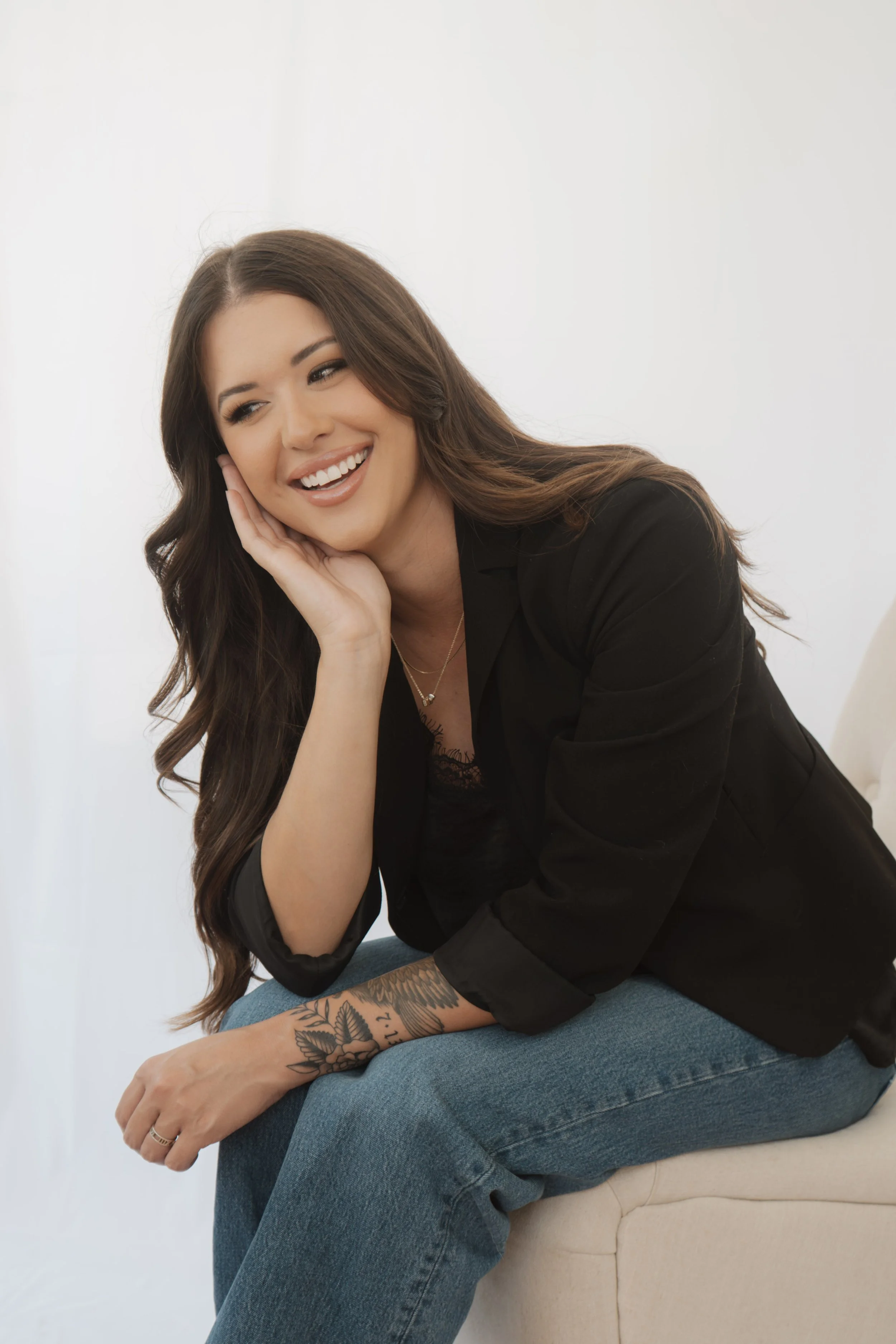 A woman with long brown hair, wearing a black blazer and blue jeans, sitting on a beige chair with her left hand resting on her face and a big smile.