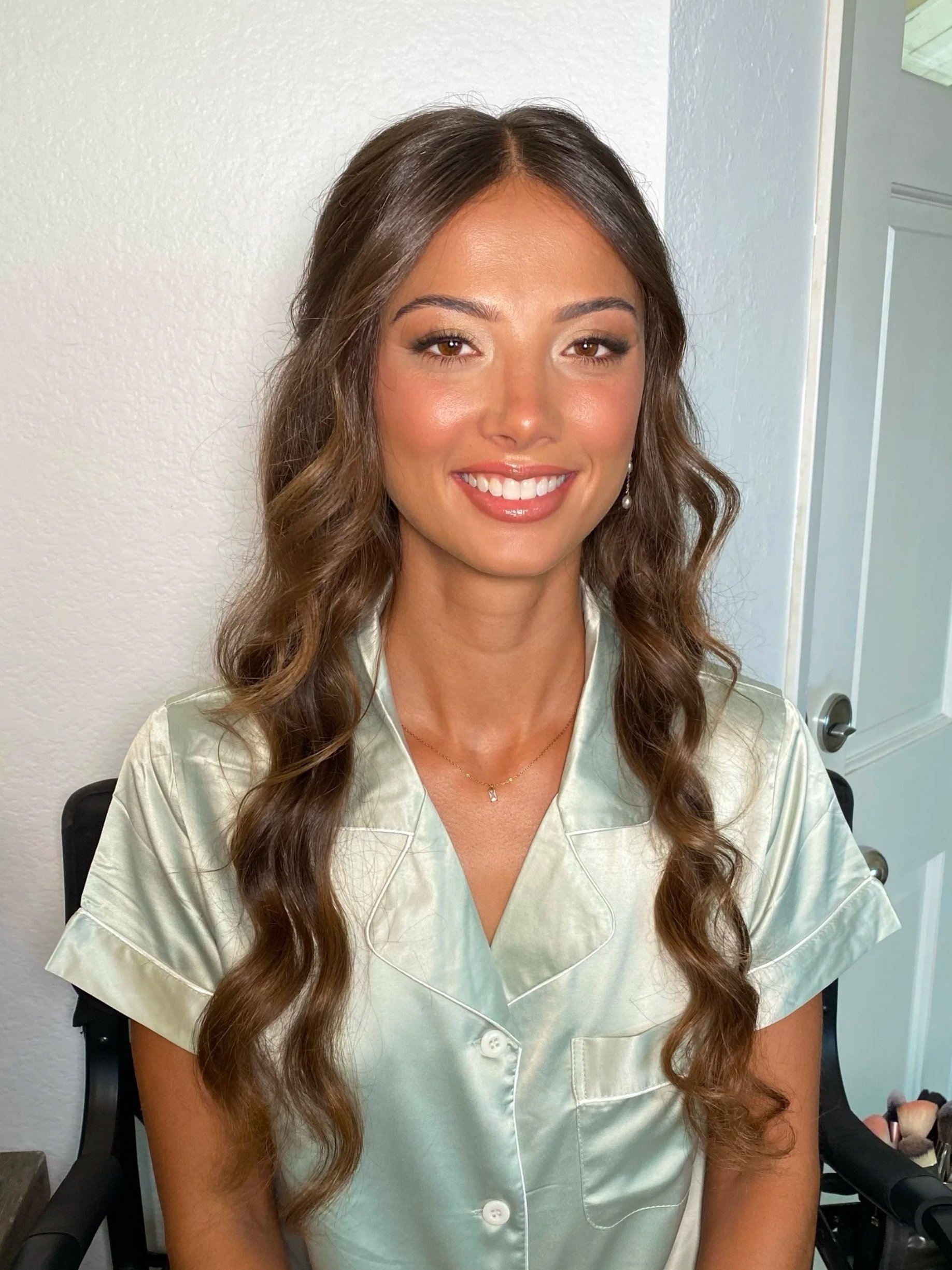 Photo of a woman with long wavy hair, wearing makeup, and a satin shirt, sitting in a room with white walls and a door visible in the background.