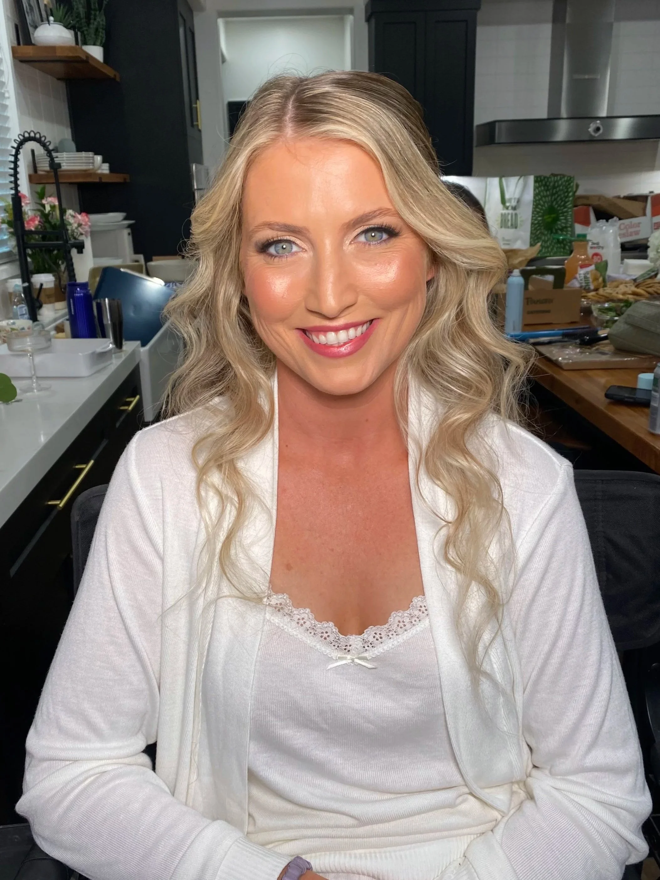 A smiling woman with blonde wavy hair in a white top sitting in a kitchen surrounded by various items on the counter and shelves.