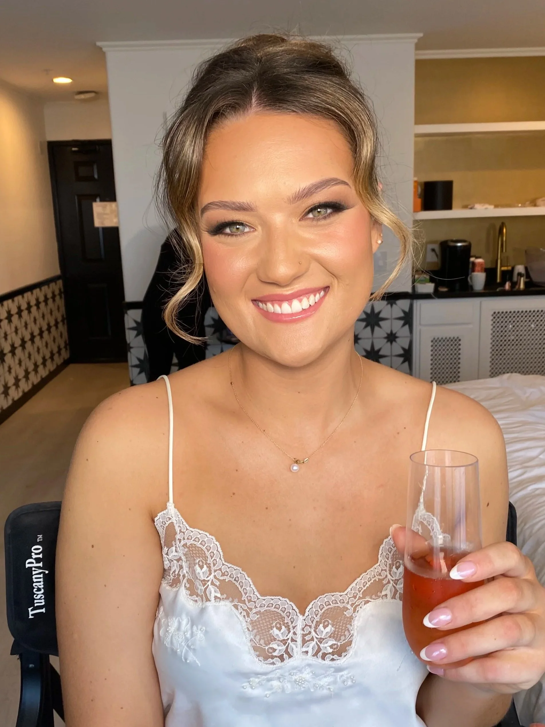 Smiling woman in a white lace dress holding a glass of rosé wine in a hotel room.