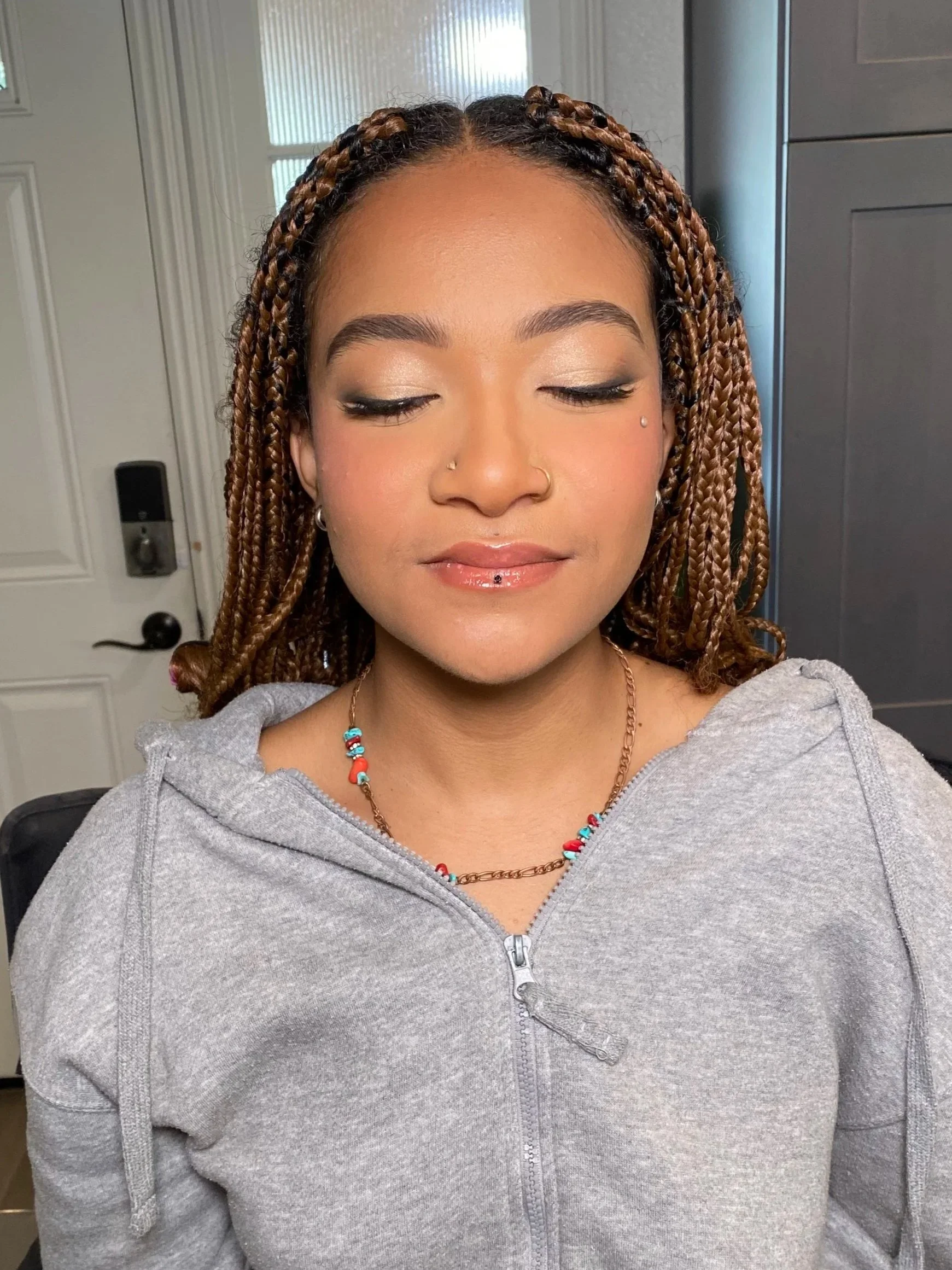 A woman with braided hair and nose piercings, wearing a gray zip-up hoodie, with her eyes closed and wearing subtle makeup and a colorful beaded necklace.