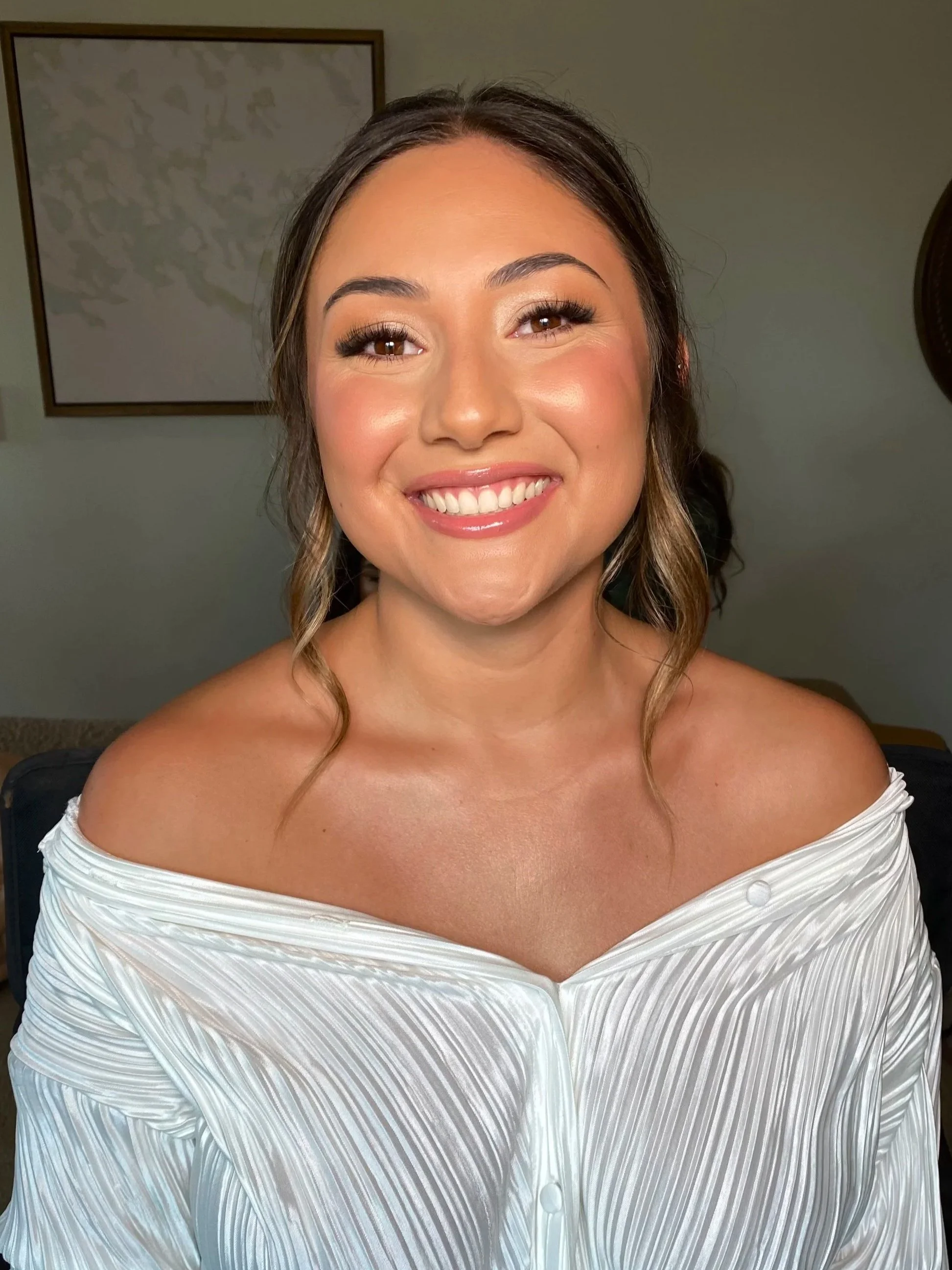 A woman with dark hair styled in loose waves, wearing makeup, and smiling broadly, and a white off-the-shoulder dress with textured stripes and buttons.