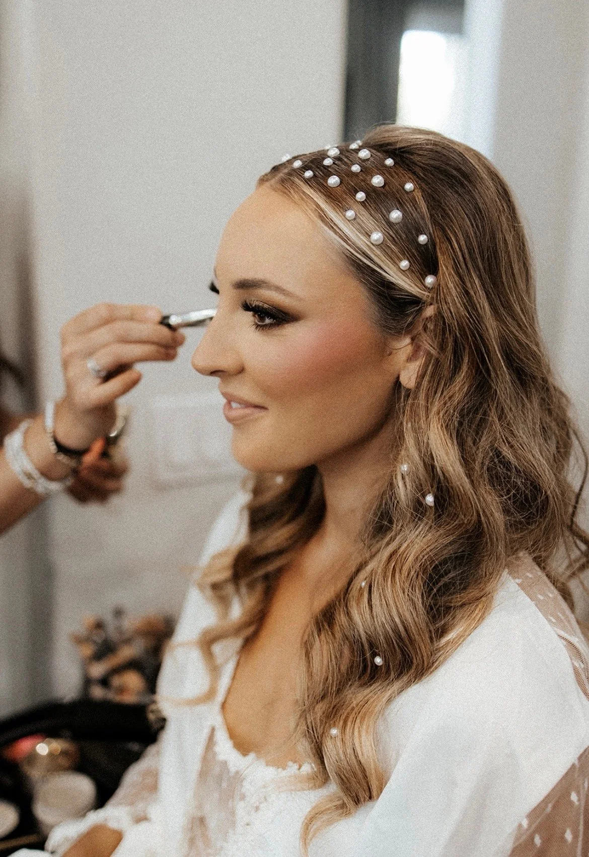 A woman with long wavy hair, adorned with pearl hair accessories, is getting her makeup done by a makeup artist.