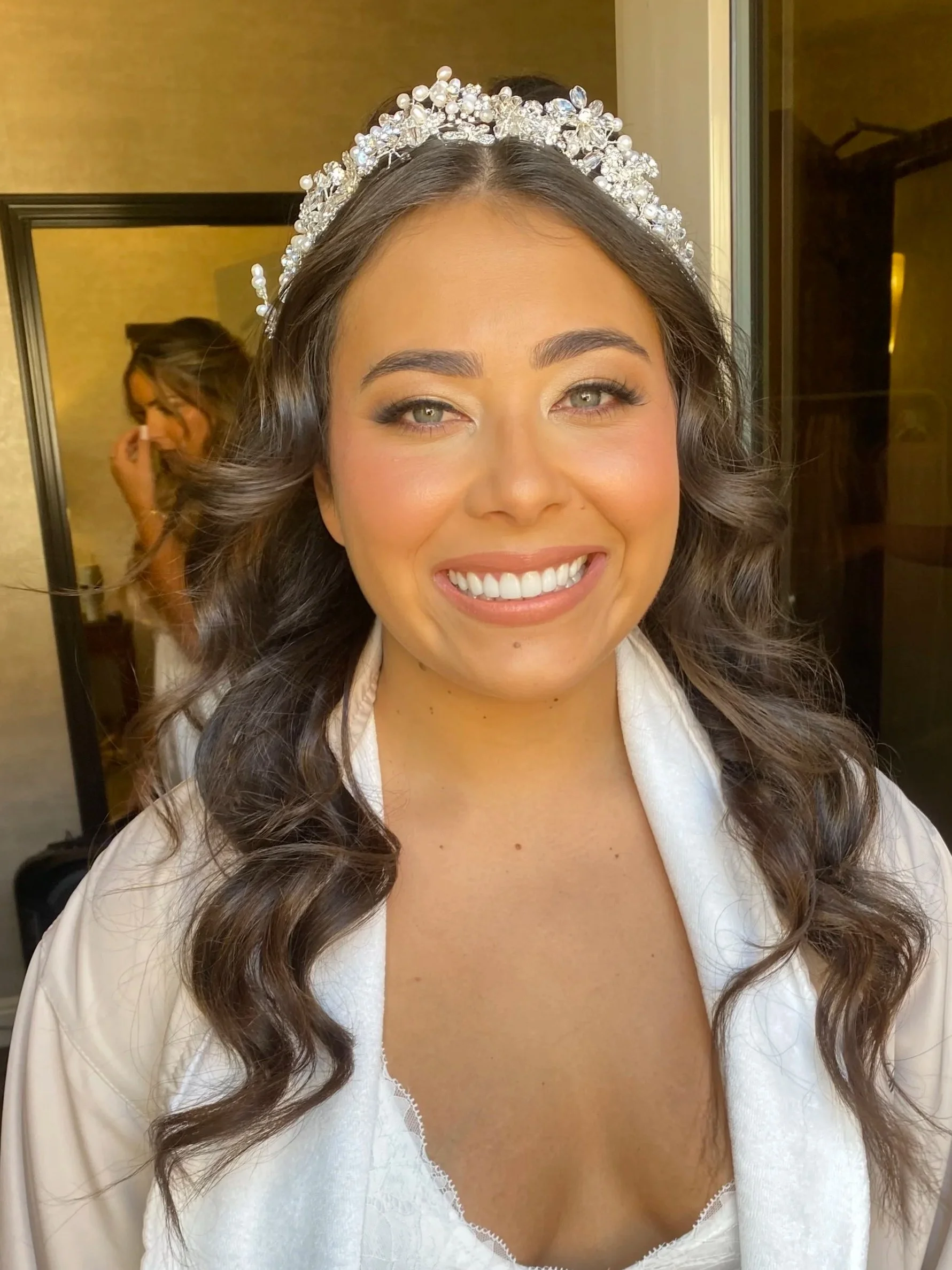 Close-up of a smiling woman with long, wavy brown hair wearing a decorative headband with pearls and rhinestones, in a room with a mirror and gold-colored walls.