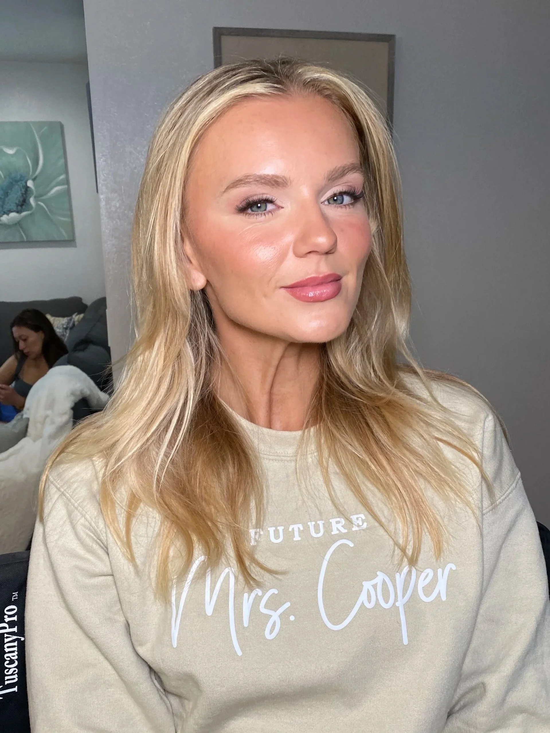 A woman with long blonde hair and light makeup, wearing a beige sweatshirt with white text, standing indoors with a gray wall and a painting of a flower behind her.