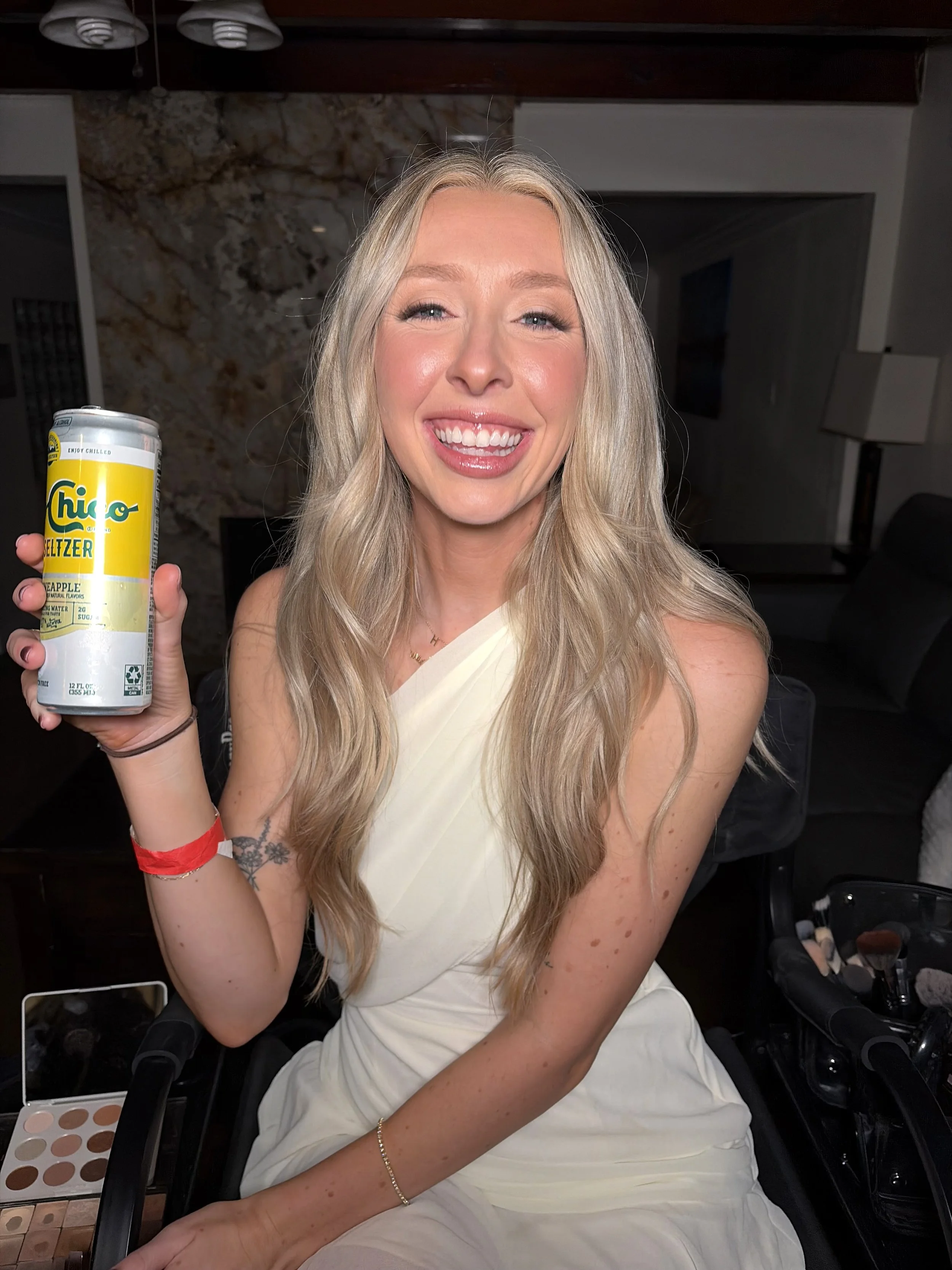 A woman with long blonde hair sitting in a makeup chair, smiling and holding a can of Chupa Chups Seltzer. She is wearing a white dress and has makeup products and brushes on a table next to her.