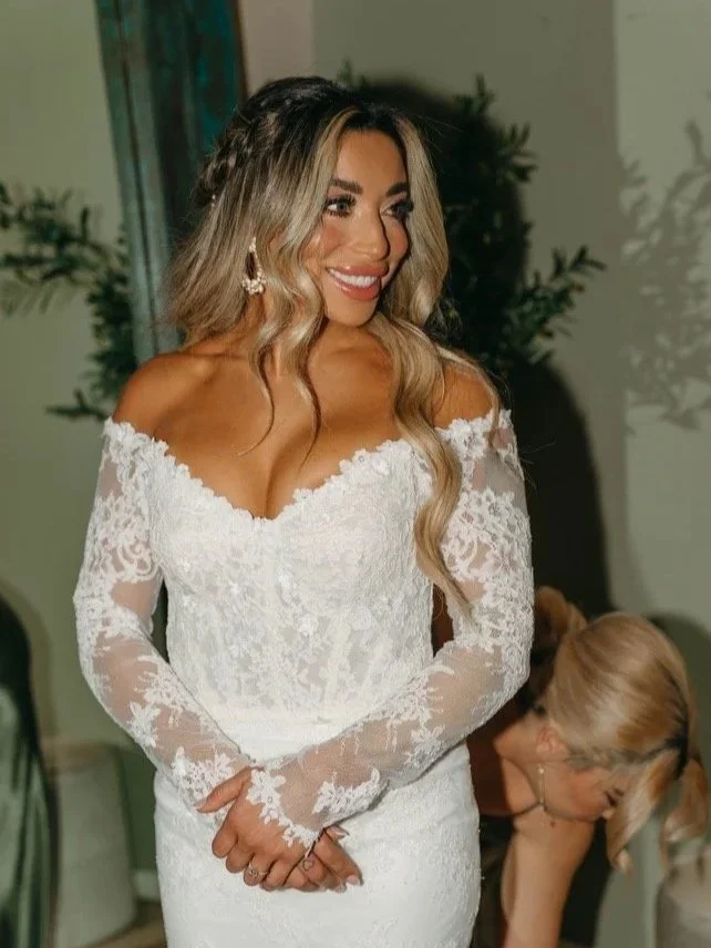 A woman in a white lace wedding dress smiling, with long wavy blonde hair and earrings, standing indoors.