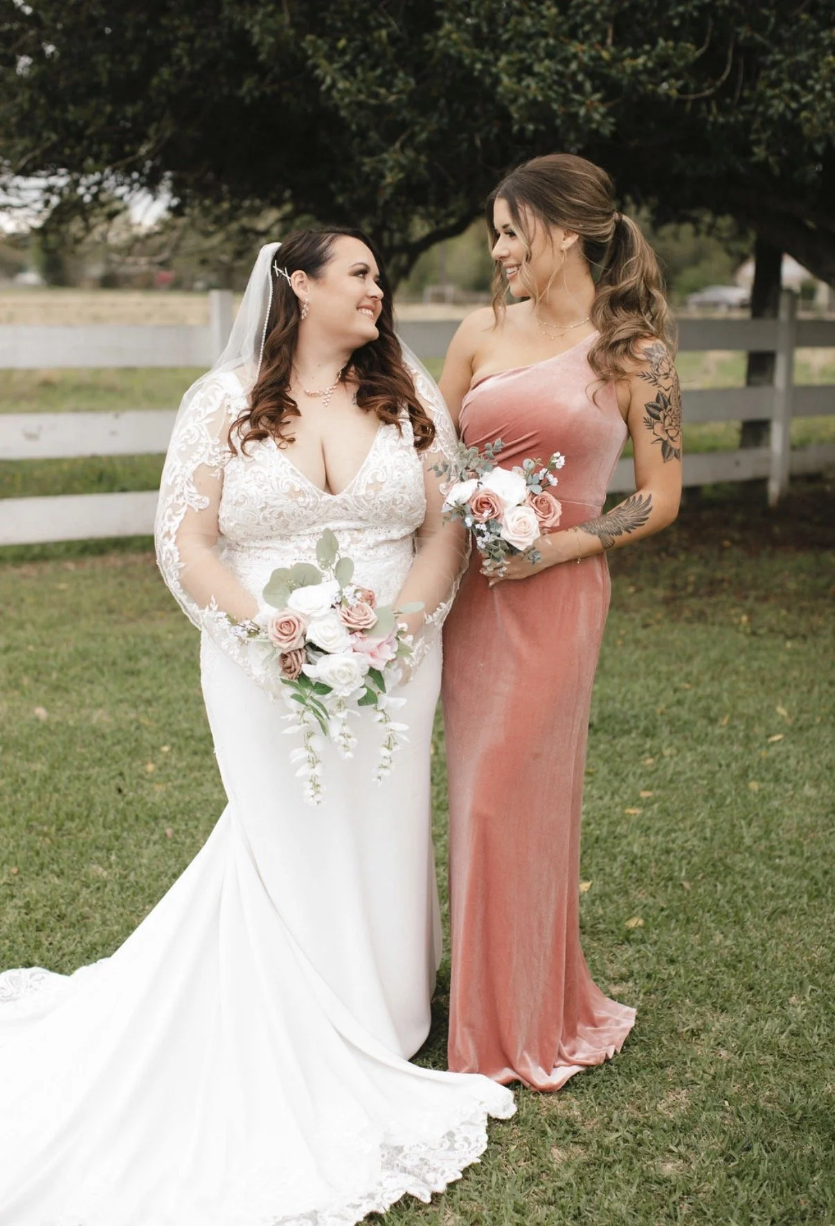 Two women standing outdoors in front of a white fence and trees, smiling at each other, holding bouquets of pink and white roses, one in a wedding dress and the other in a pink gown.