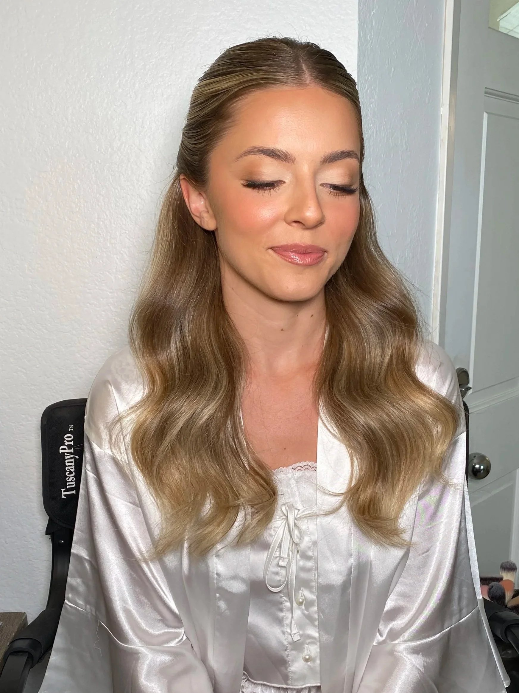 A woman with long, wavy blonde hair wearing a white satin pajama top, sitting in a wheelchair with a black backrest labeled "TuscanYPro." She has her eyes closed and a gentle smile.