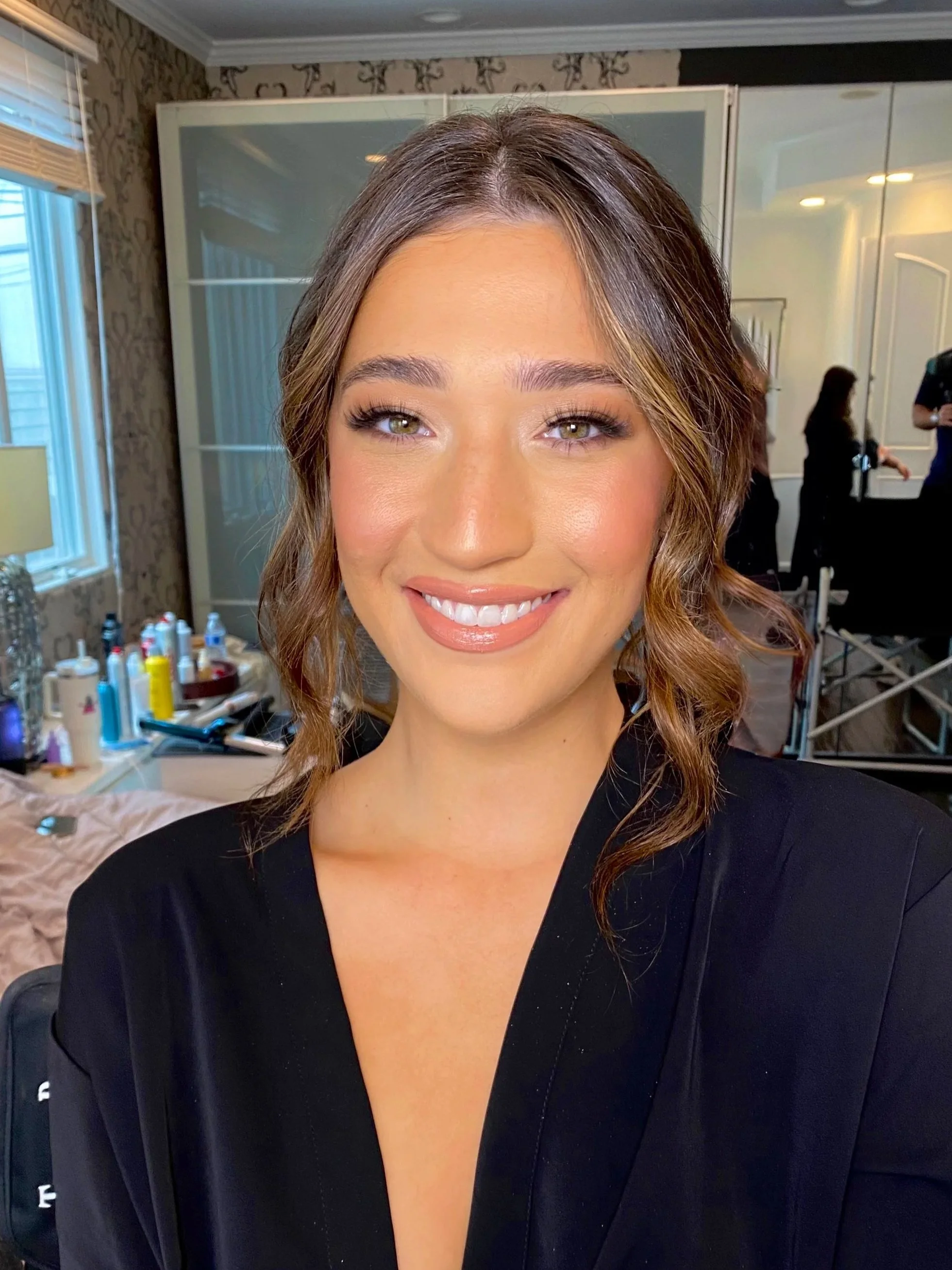 A woman smiling with brown wavy hair, wearing makeup, in a room with a mirror and shelves with beauty products in the background.