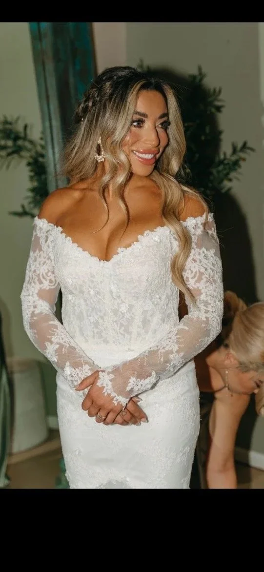 A woman in a white lace wedding dress with off-the-shoulder long sleeves, smiling, with blonde wavy hair, earrings, and a braid in her hair.