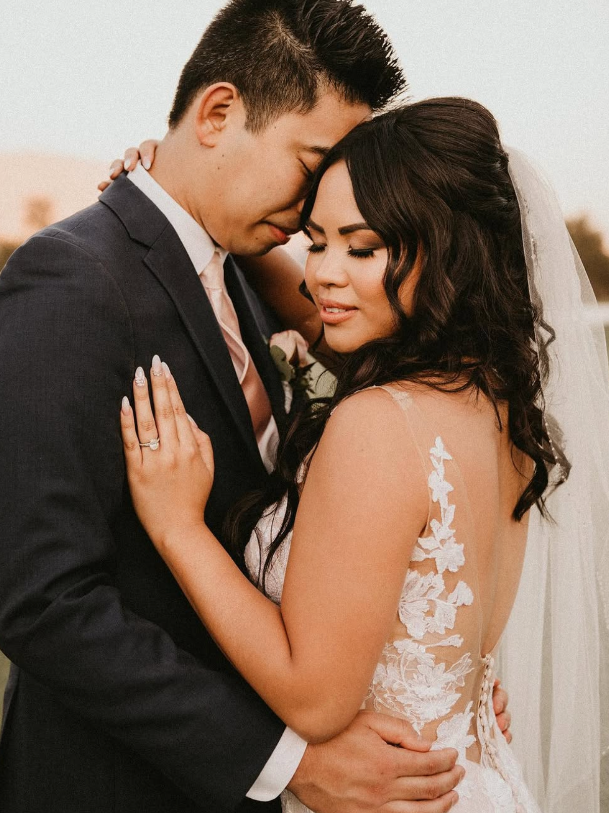 A newlywed couple is embracing each other, with their foreheads touching and eyes closed, outside during sunset. The groom is wearing a dark suit and the bride is in a white wedding dress with floral lace details.