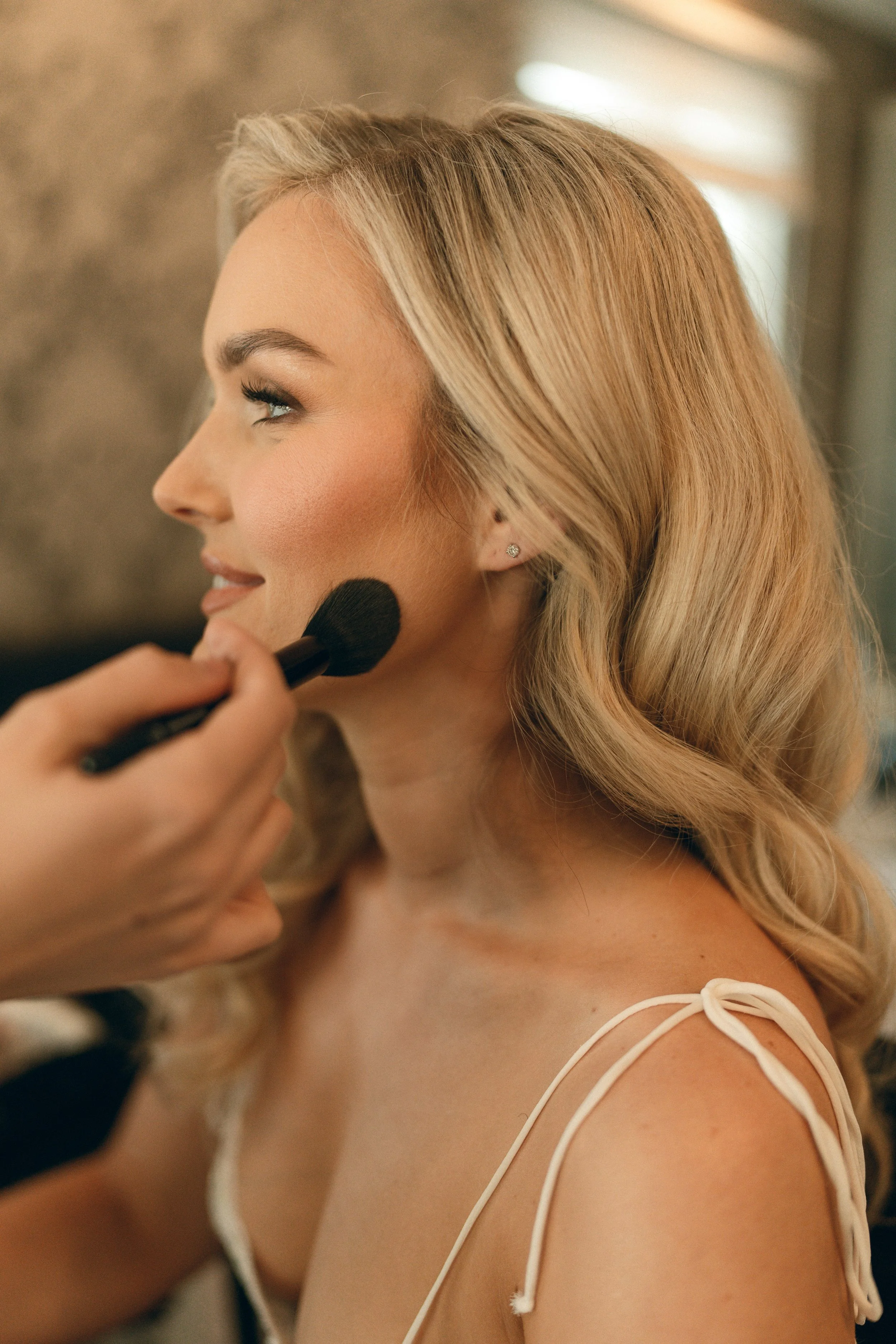 A woman with blonde hair getting her makeup done, with makeup brush near her cheek.