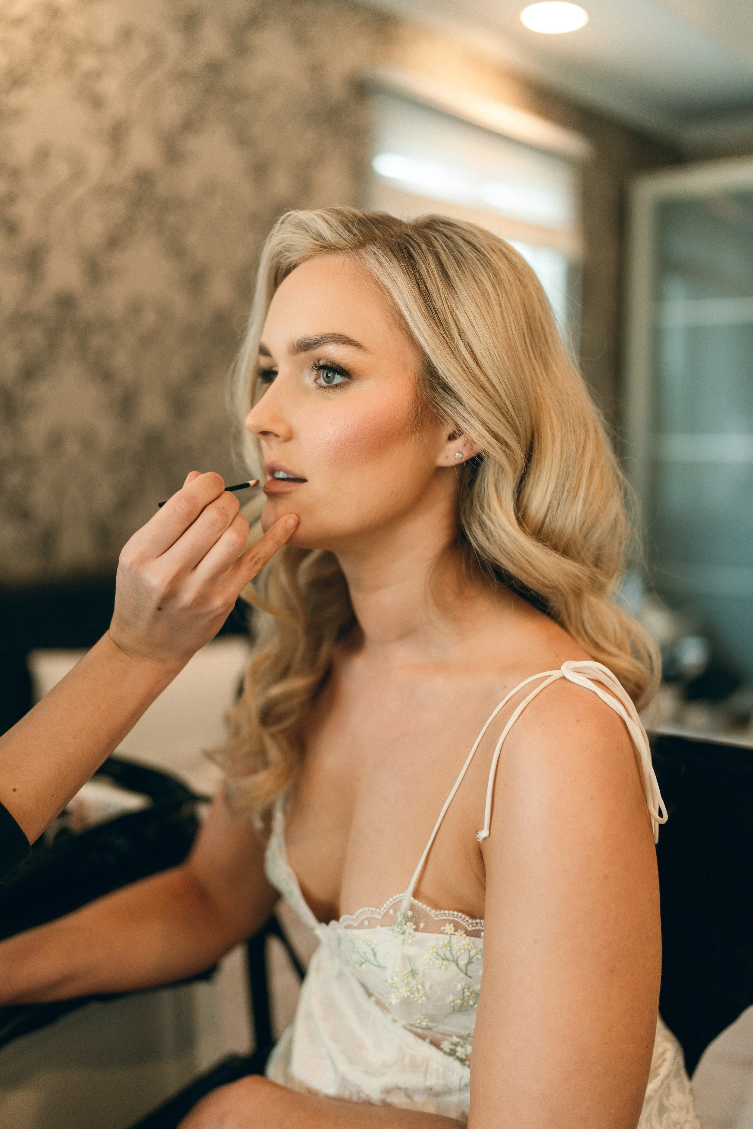 A woman with blonde hair getting her makeup done by a makeup artist, sitting in a well-lit room with a stone wall background and a window.