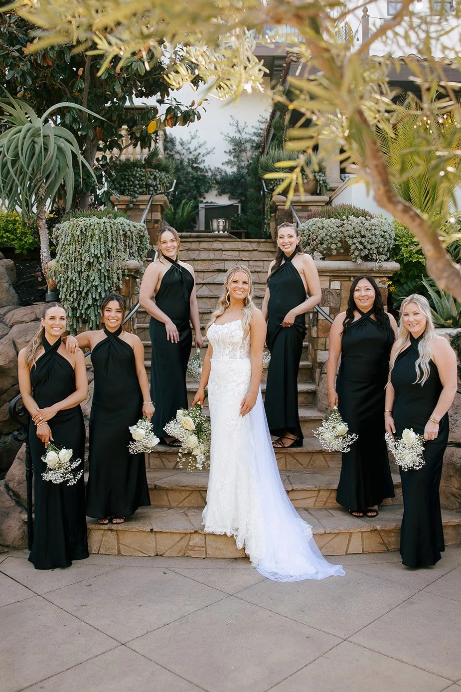 A bride in a white wedding dress surrounded by six bridesmaids in black dresses, standing on outdoor stone stairs with greenery and plants in the background.