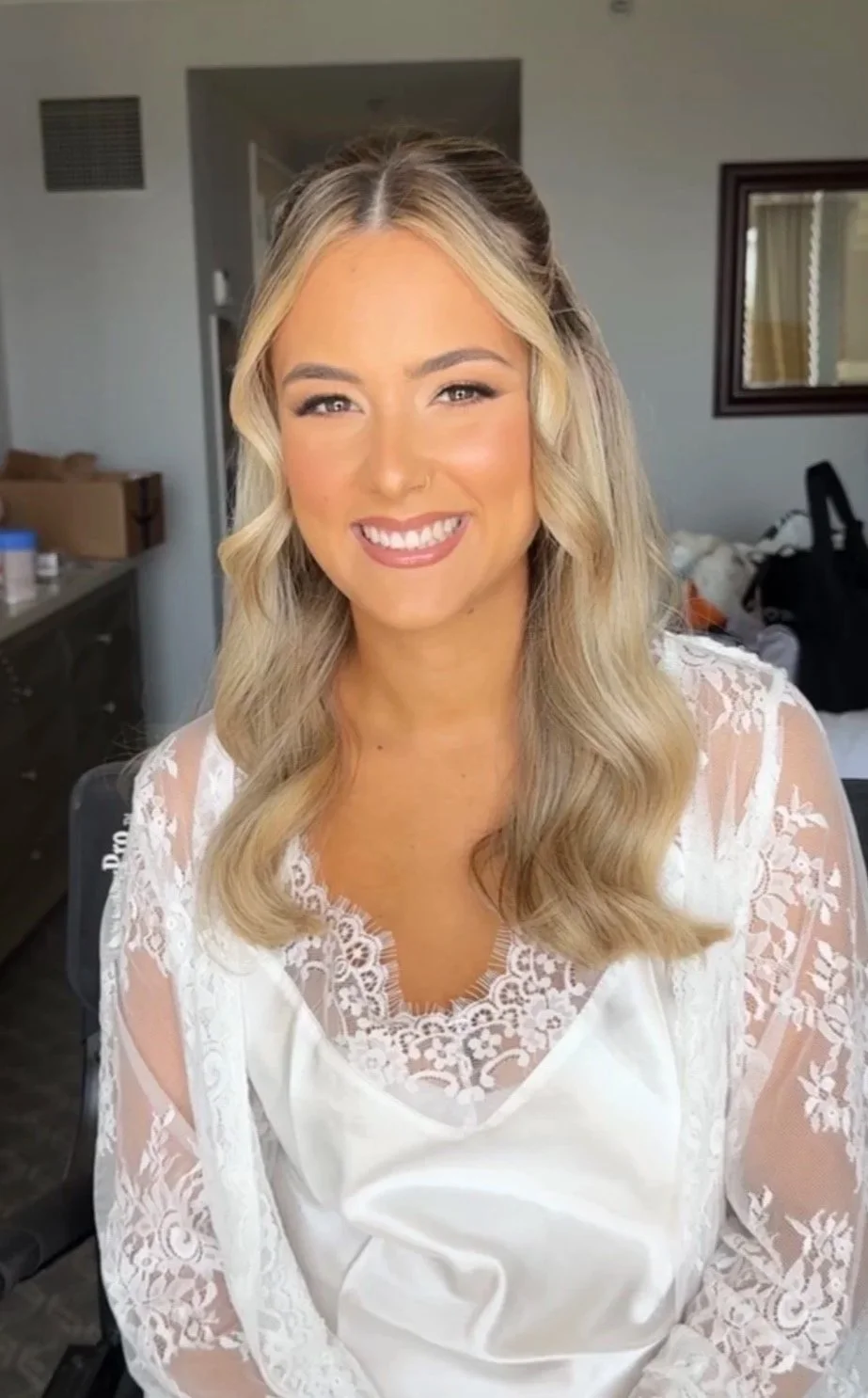 A smiling woman with wavy blonde hair, wearing a white lace top, indoors in a well-lit room.