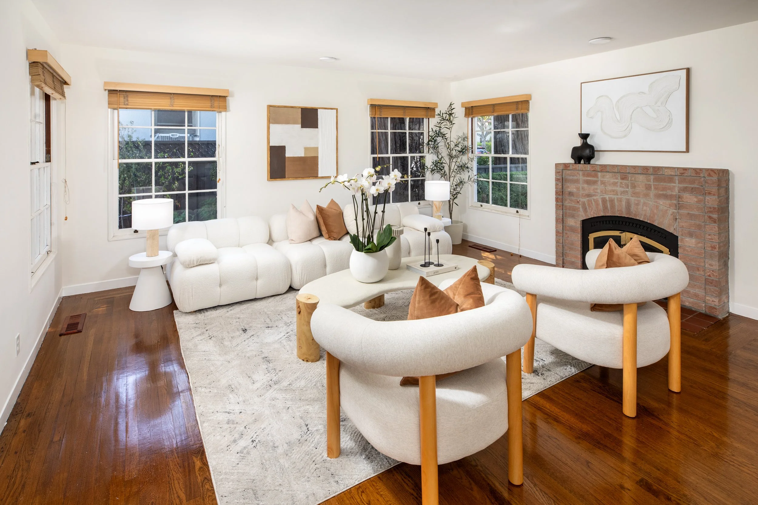 Living room with white sofas and armchairs, wooden flooring, a fireplace with artwork and a black vase, windows with wooden blinds, and a white area rug.