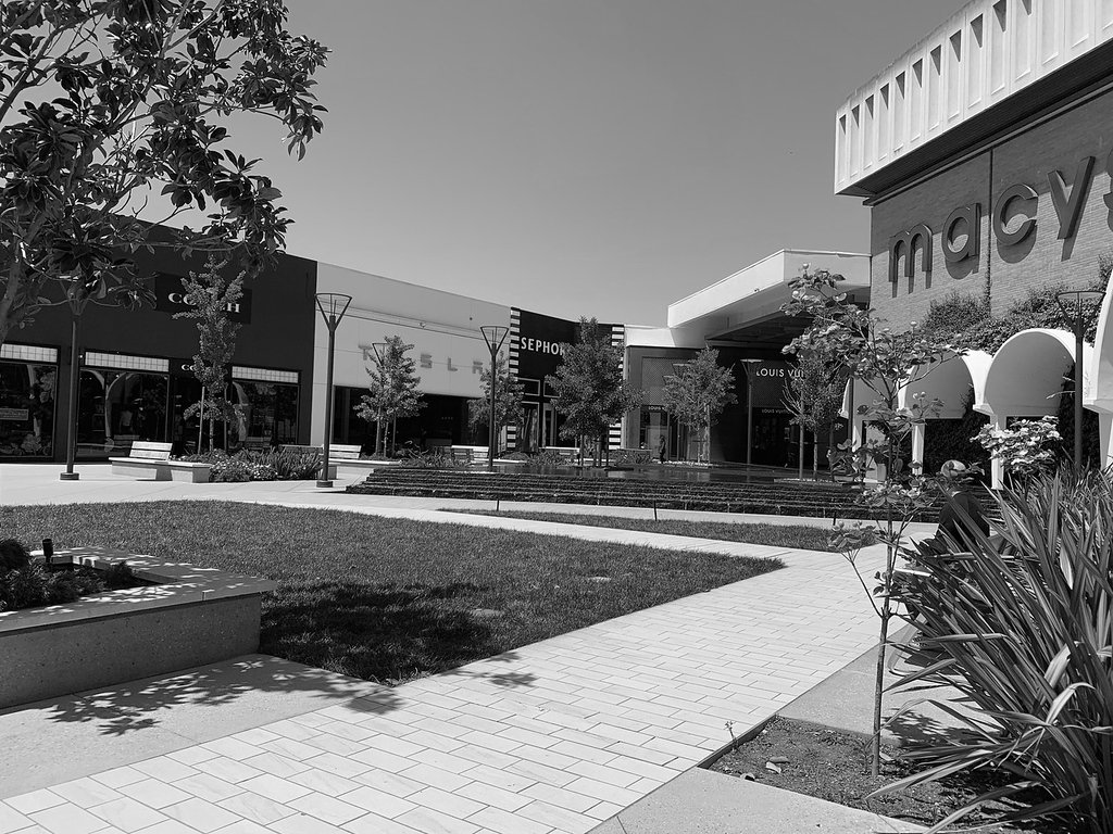 A black and white photo of an outdoor shopping mall, showing stores like Macy's and Sephora, with benches, trees, and landscaped areas.